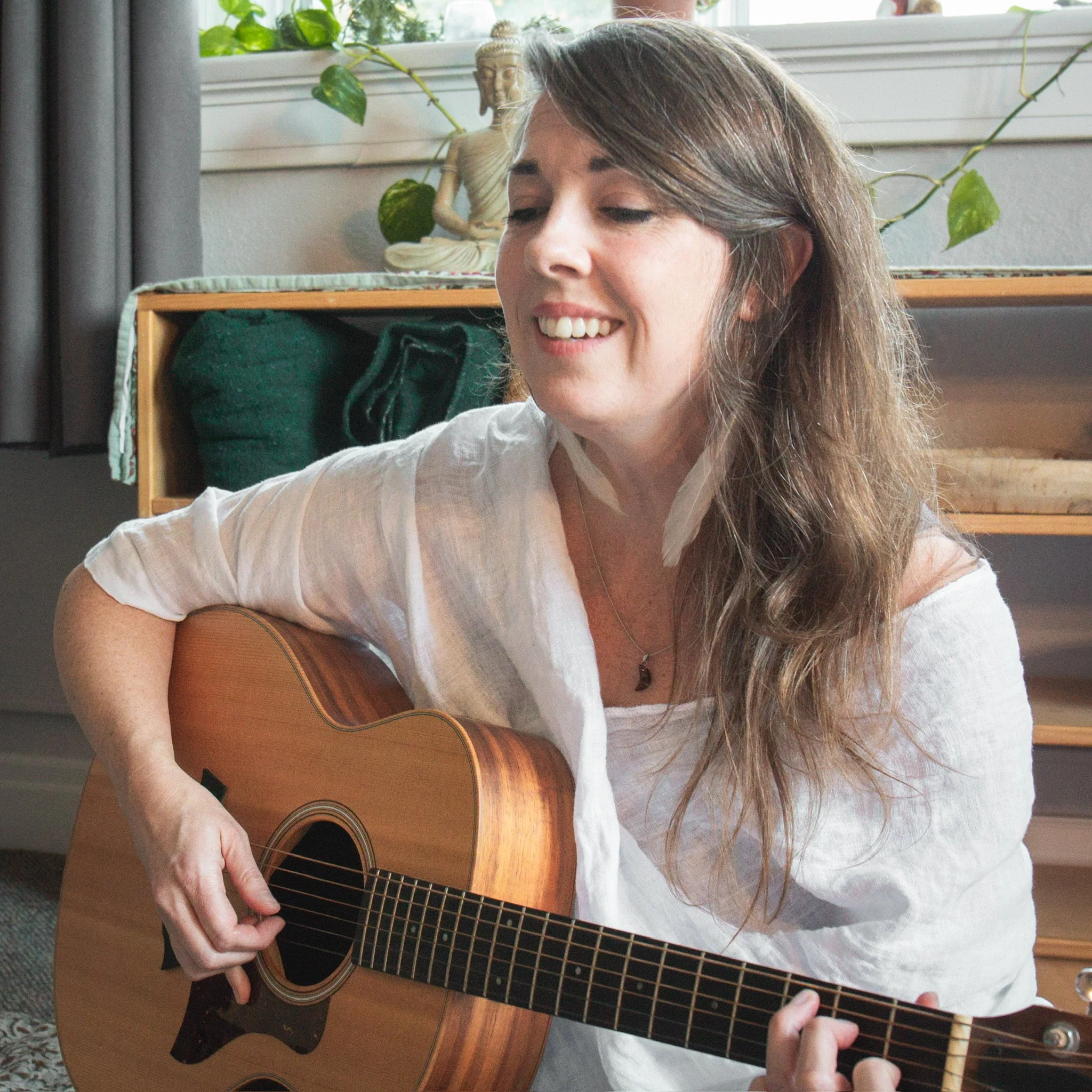 A woman playing an acoustic guitar indoors, smiling, with a Buddha statue and houseplants in the background.
