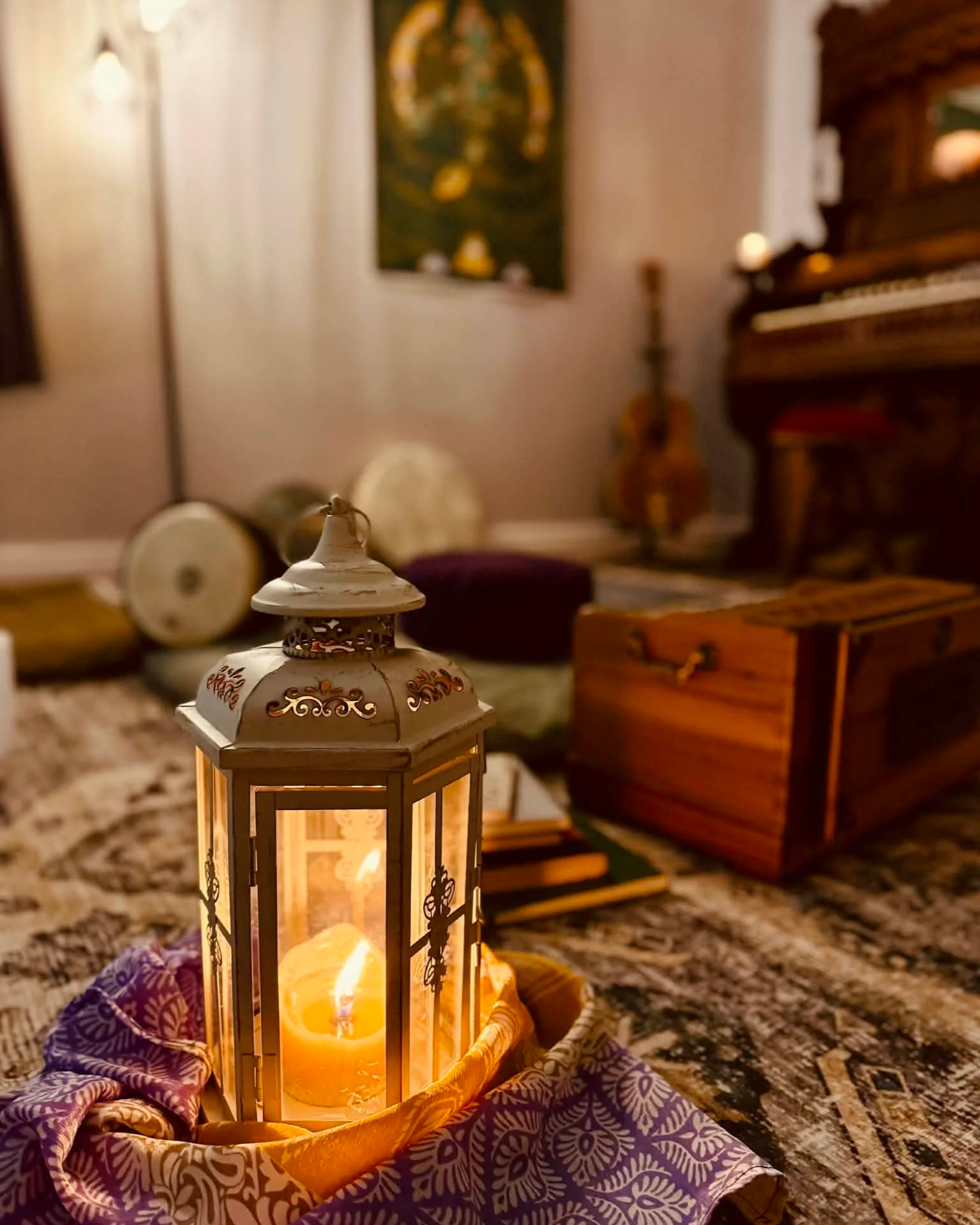 A lit candle inside a decorative lantern on a patterned cloth, with musical drums, a guitar, a wooden chest, and a bookshelf in the background.