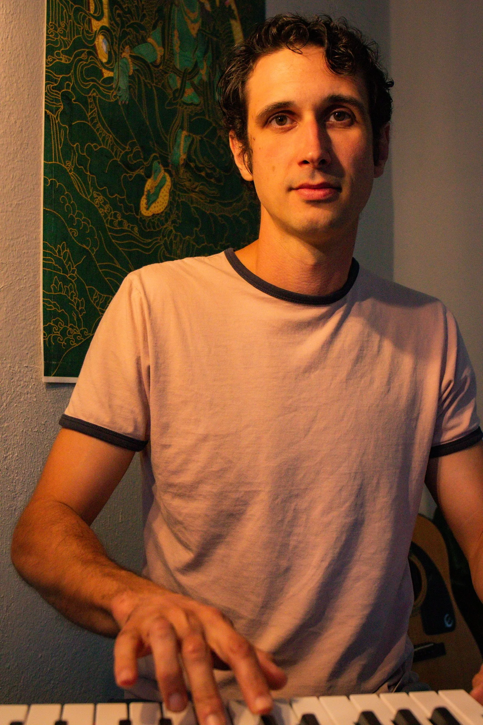 A young man with dark, curly hair and fair skin is sitting at a keyboard piano, looking at the camera. He is wearing a light beige t-shirt with black trim around the neck and sleeves. Behind him is a decorative wall hanging with a dragon or serpent design in gold and green.