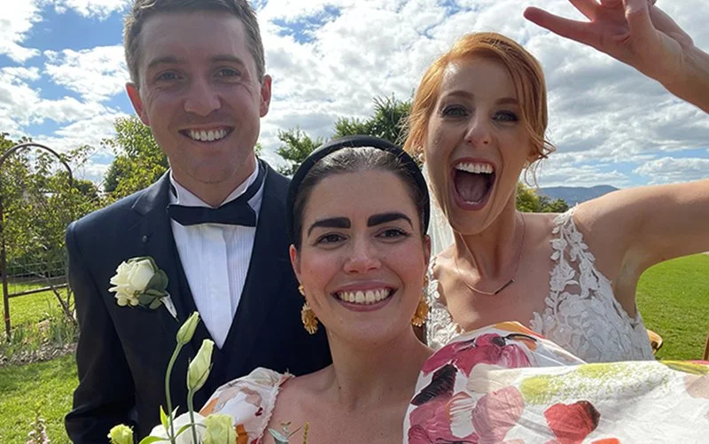 Three people at an outdoor wedding, smiling and taking a selfie. The groom is dressed in a tuxedo with a white shirt and black bow tie. The bride has red hair, is wearing a white wedding dress with floral details, and is waving. The woman in the middle has dark hair, gold earrings, and is holding a bouquet of white flowers.