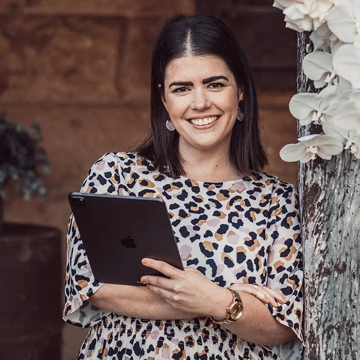 A woman with dark hair smiling and holding a tablet, standing next to a tree with white flowers.
