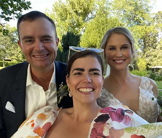 Three smiling people taking a selfie outdoors with trees in the background.