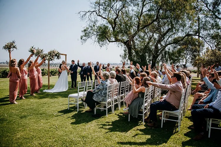 Outdoor wedding ceremony with bride, groom, bridesmaids, groomsmen, and seated guests.