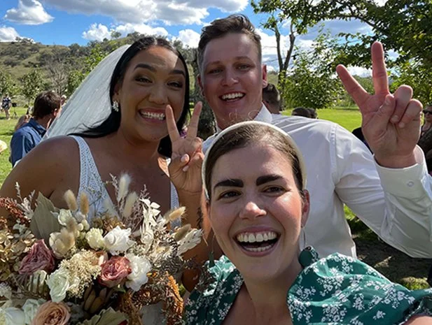 Three people at an outdoor wedding, smiling and making peace signs, with a bride holding a bouquet of flowers and trees in the background.