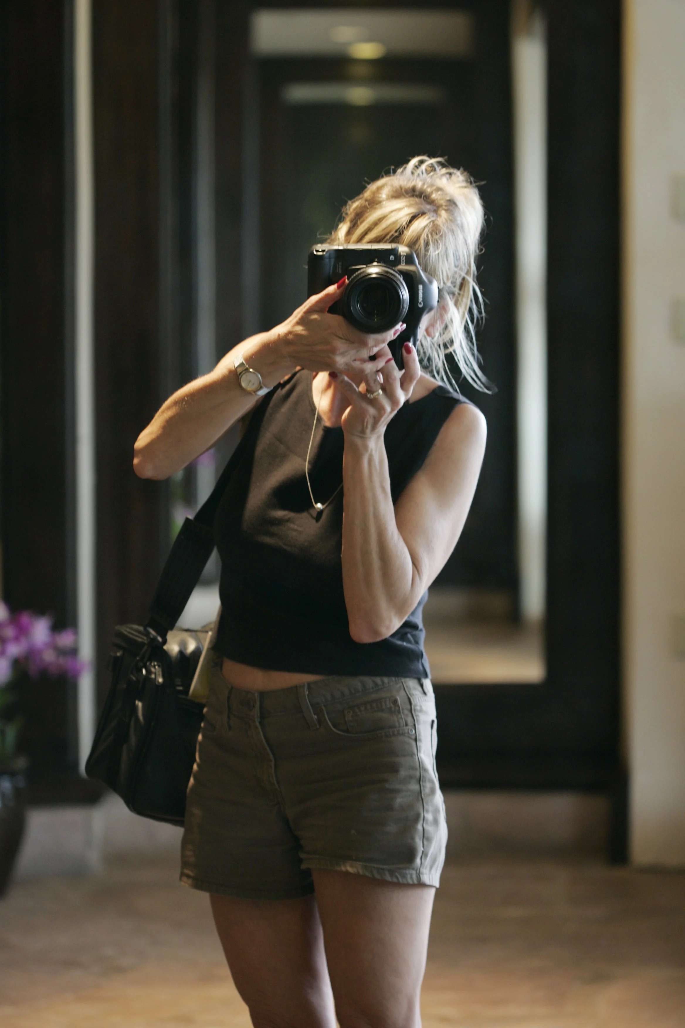 A woman with blonde hair taking a photograph in front of a mirror using a DSLR camera. She is wearing a black sleeveless top, khaki shorts, a watch, and a necklace. She has a black bag slung over her shoulder.