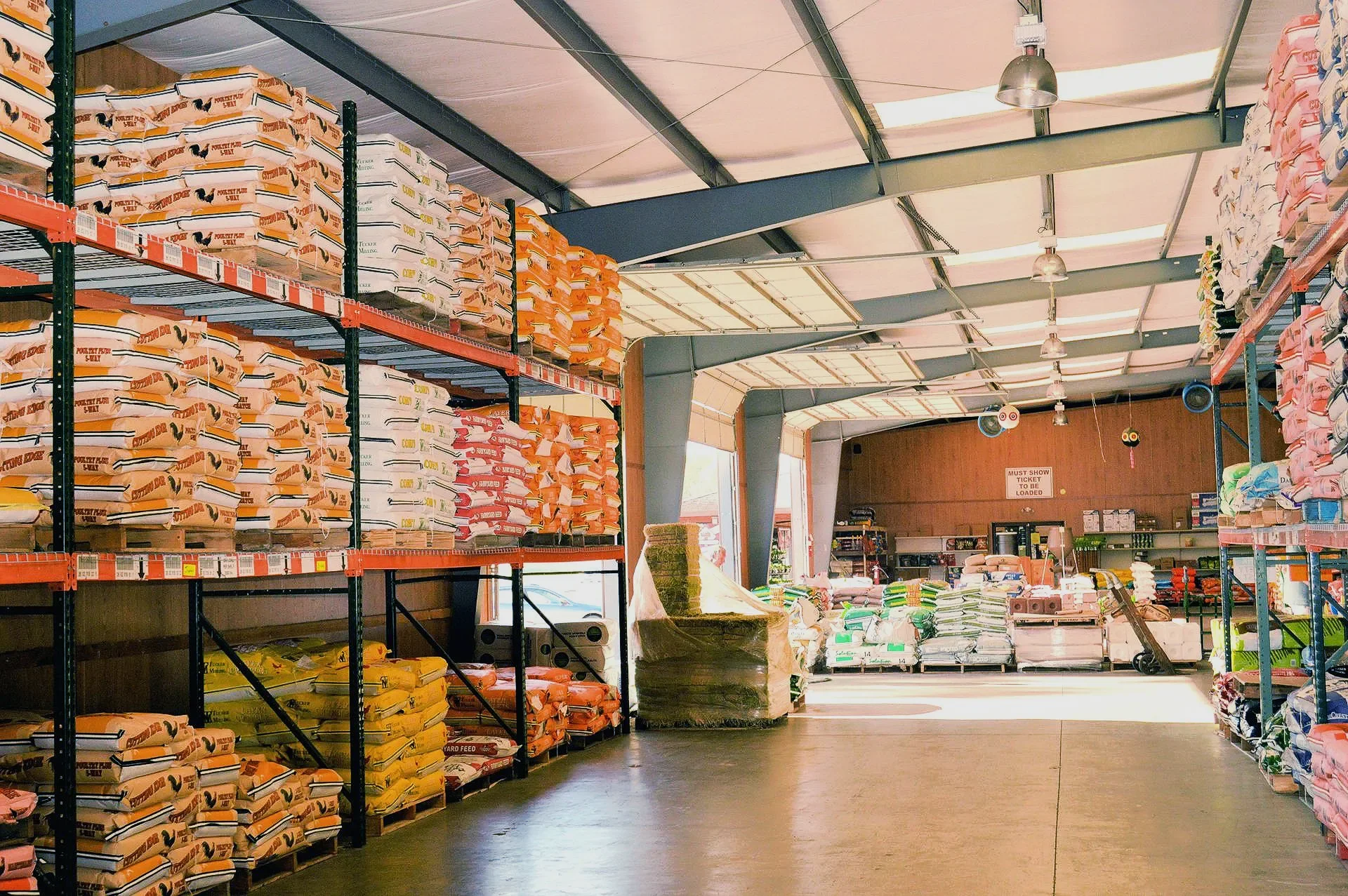 Interior of a store aisle with shelves stocked with bags of pet or animal feed, illuminated by hanging lights, with pallets and a chair near the back.