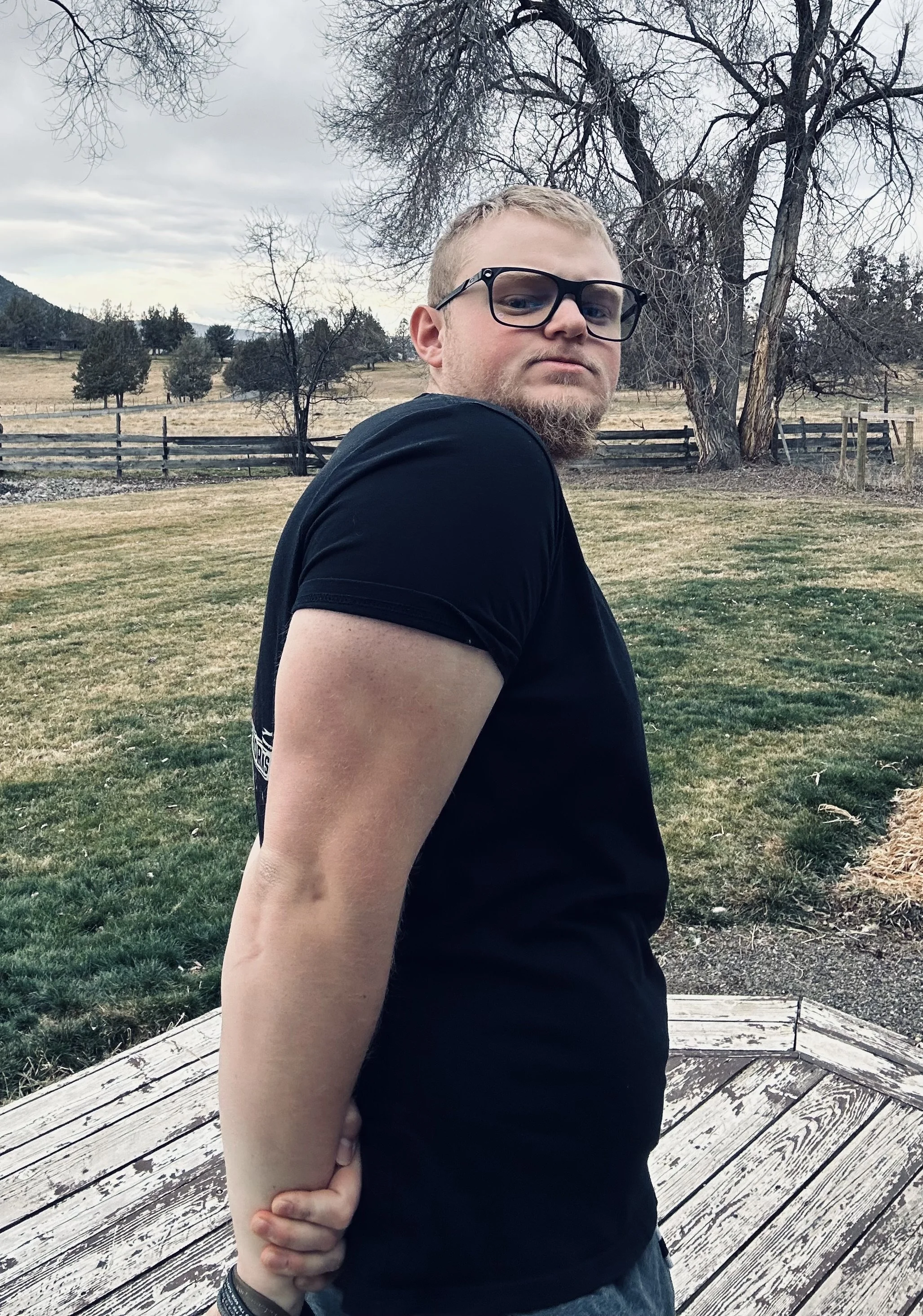 Young man with glasses and beard wearing a black t-shirt, standing outdoors on a wooden platform with an overcast sky, leafless trees, grassy field, and wooden fence in the background.