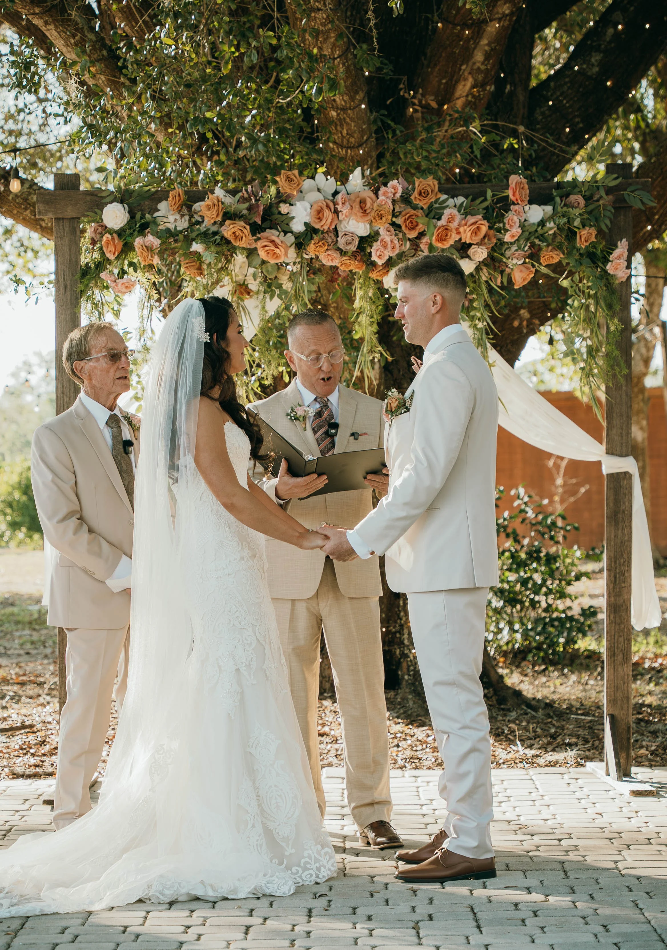 A couple gets married outdoors under a floral arch decorated with pink and white roses. The bride and groom hold hands while an officiant, standing between them, reads vows or words. Two older men stand nearby, possibly family members, in cream-color
