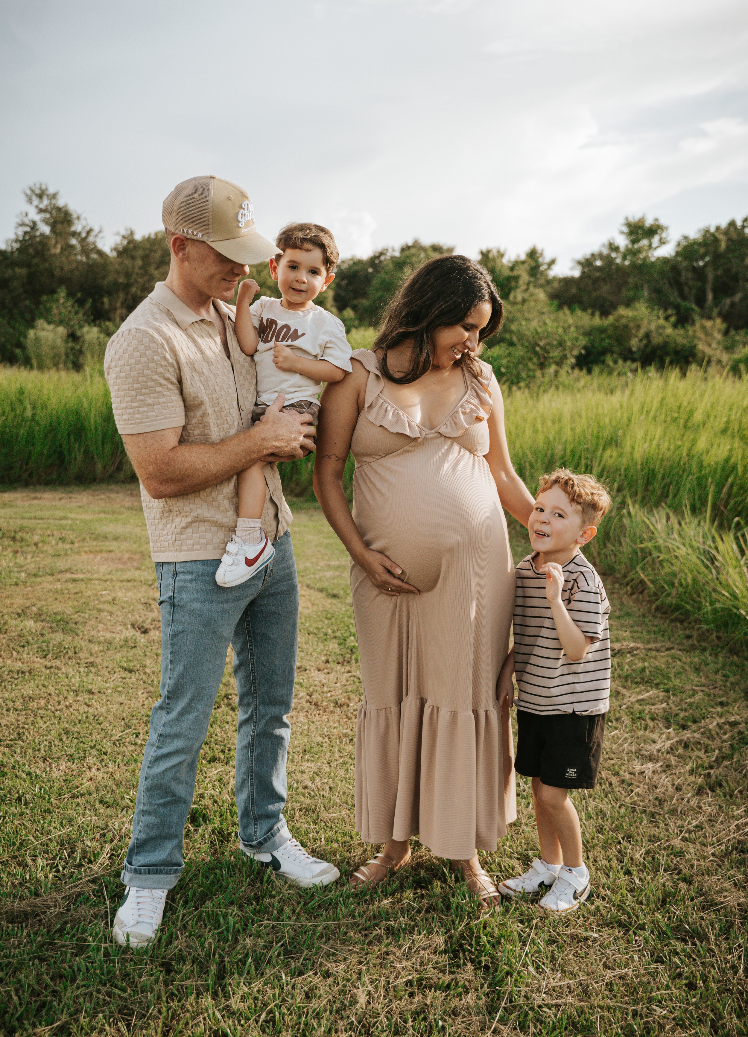 Family of four, including pregnant woman, standing on grass field with tall green grass and trees in the background on a cloudy day, with the woman smiling and touching her belly while the children look at her and the man holding the younger child.