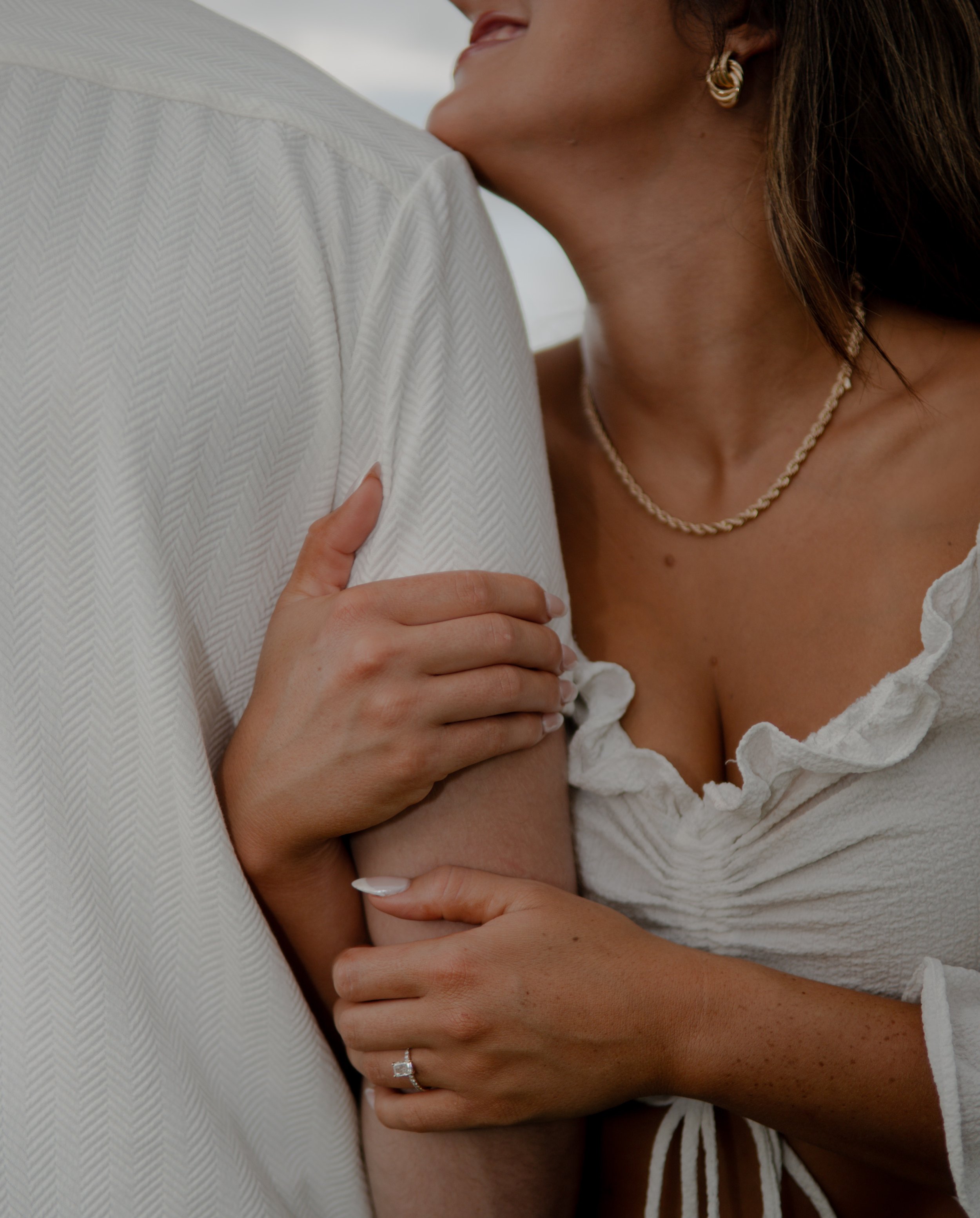 Close-up of a couple embracing, focusing on the woman's hands on the man's arm, the woman's face turned towards the man's shoulder, wearing jewelry and a white top with ruffles.