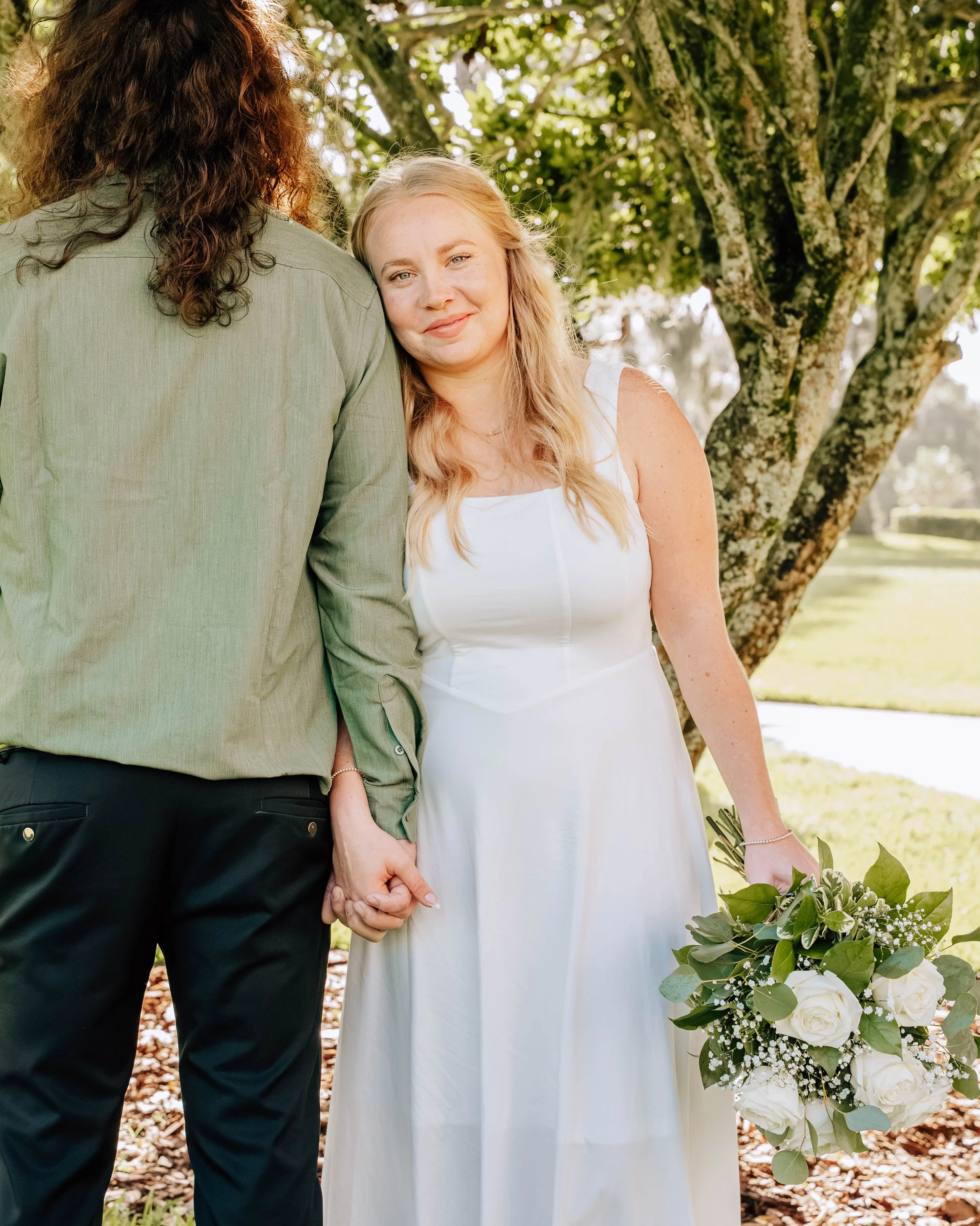 A woman in a white dress holding a bouquet of white roses and greenery, standing outdoors by a tree with a man, whose face is not visible, holding her hand.
