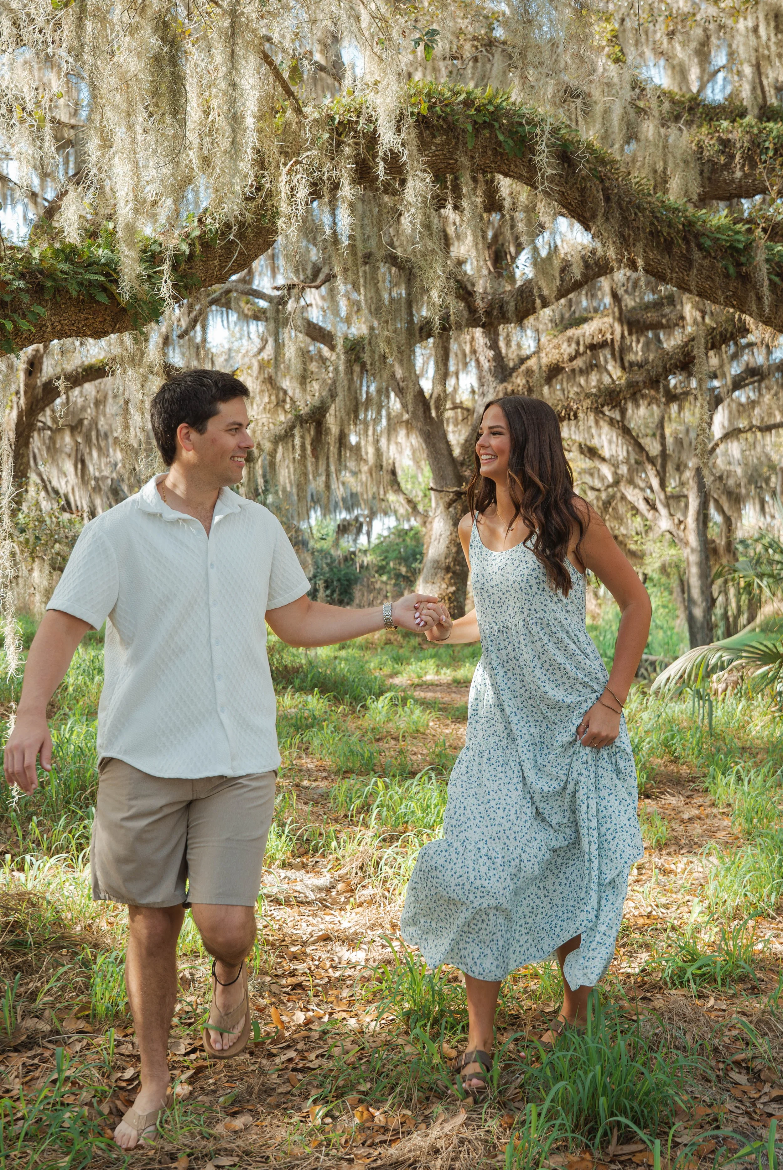 A happy couple holding hands and running through a wooded area with hanging moss and tall trees.