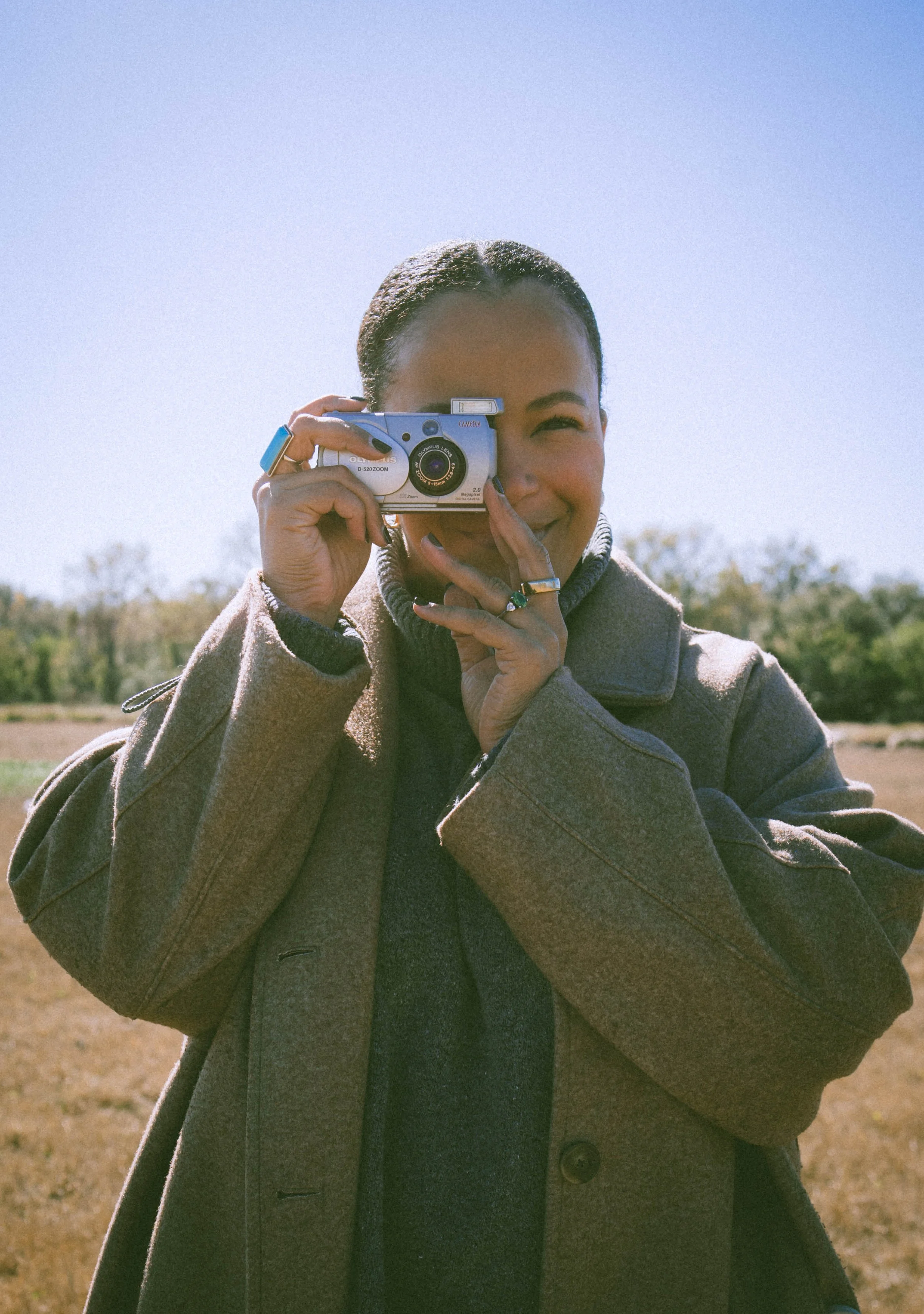 A woman standing outdoors taking a picture with a vintage camera, wearing a gray coat, with a sunny sky and trees in the background.