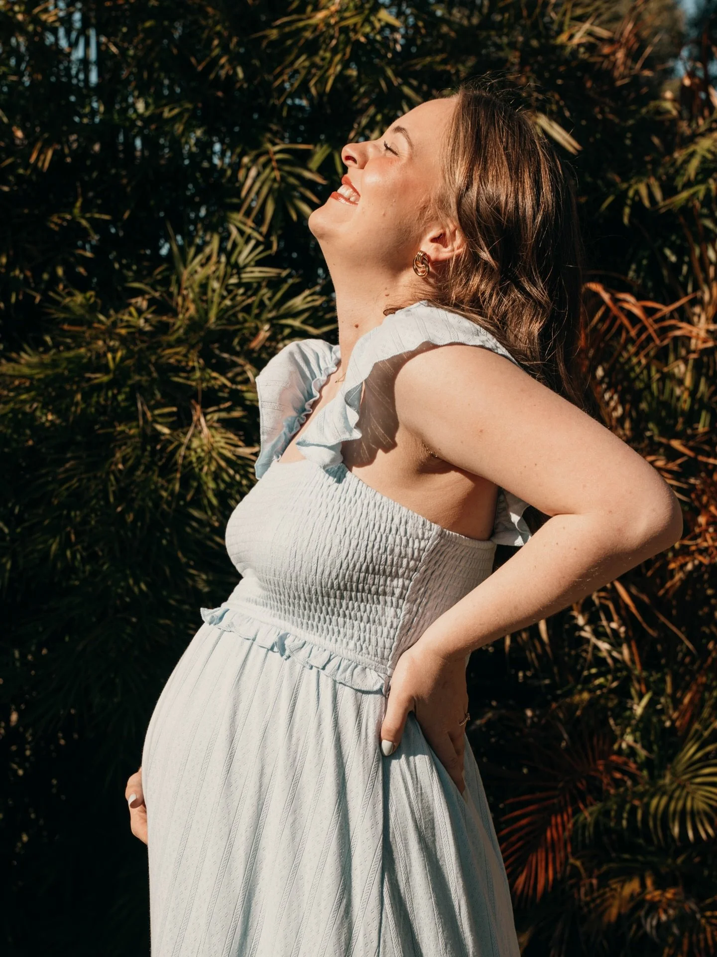 A pregnant woman smiling and looking up outdoors with lush green foliage in the background, wearing a light-colored dress.