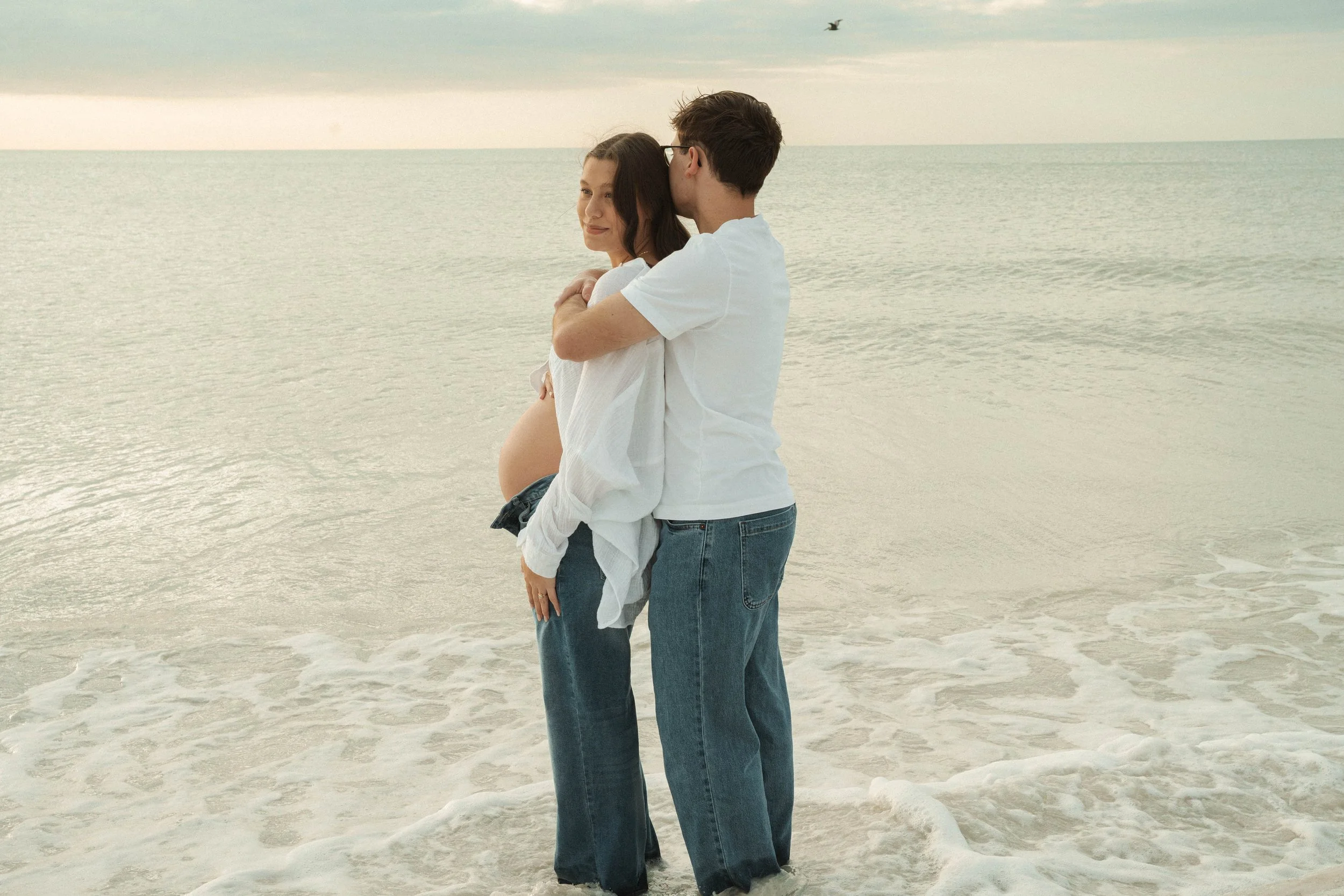 A pregnant woman and a man are standing on the beach by the ocean, with the man hugging the woman from behind, both wearing casual clothes and enjoying the moment.