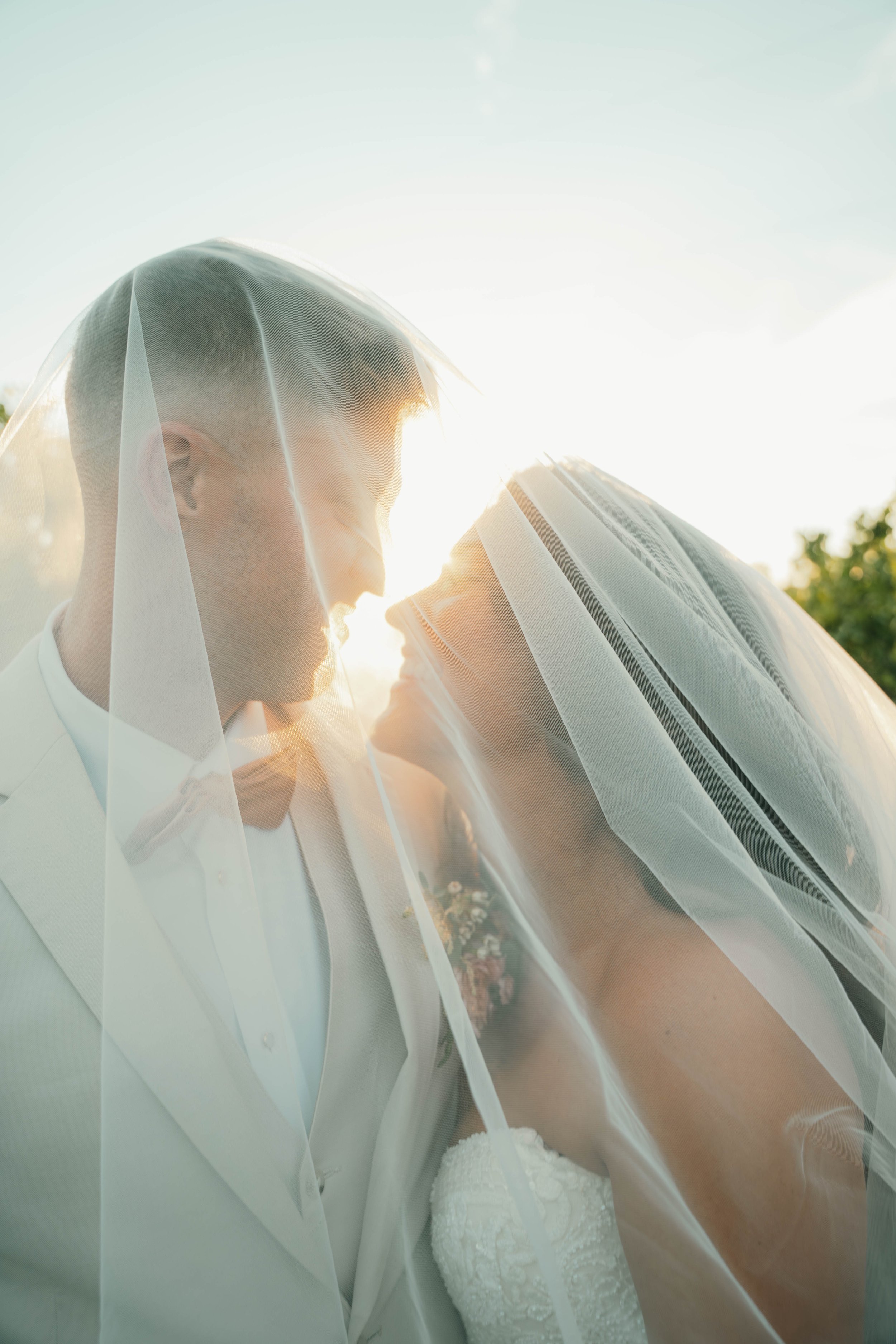 A newlywed couple standing close under a sheer veil, with the sun shining brightly behind them, creating a romantic glow.
