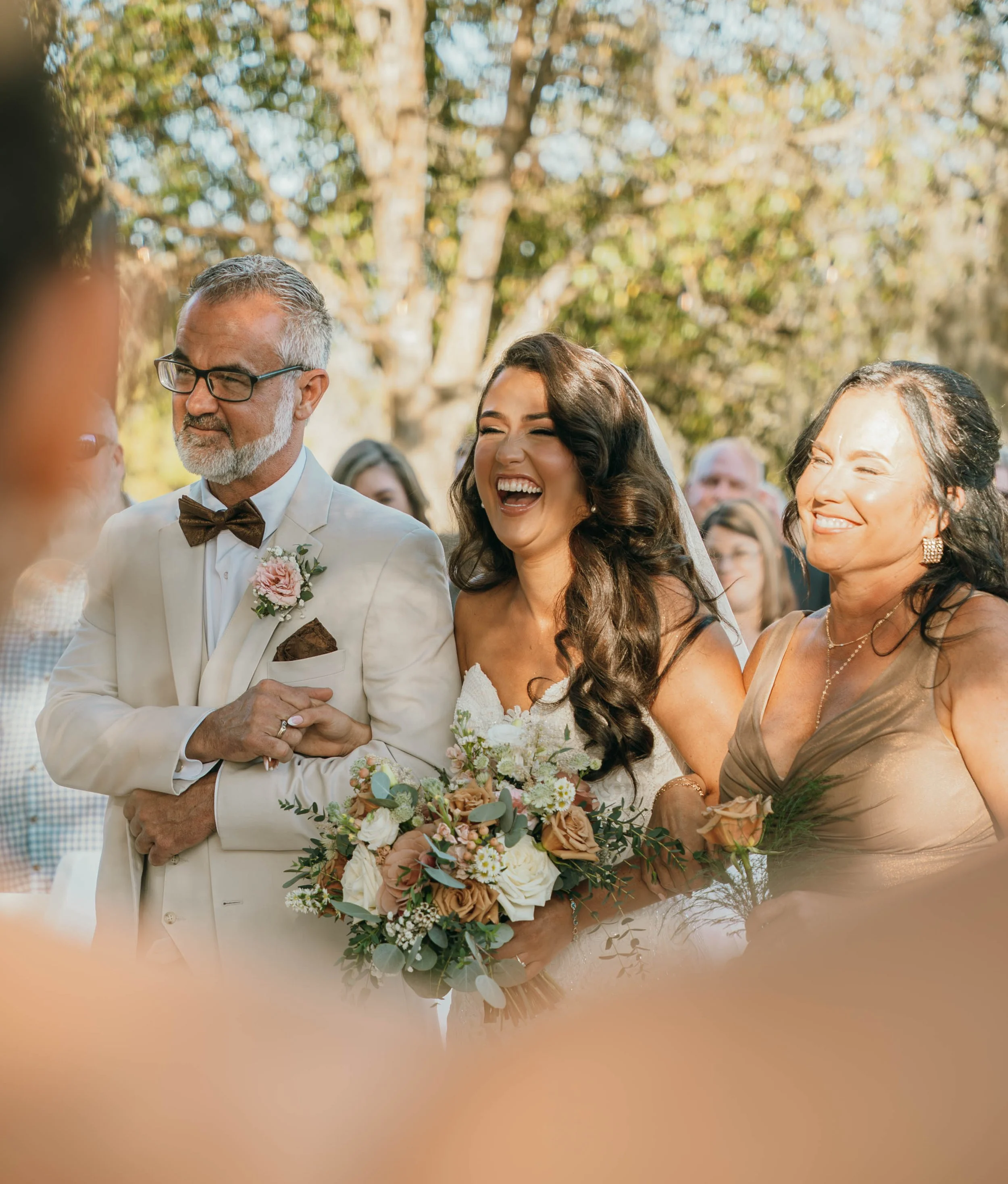 A bride laughing and holding a bouquet of flowers standing between an older man in a white suit with glasses and a woman in a gold dress at an outdoor wedding ceremony.