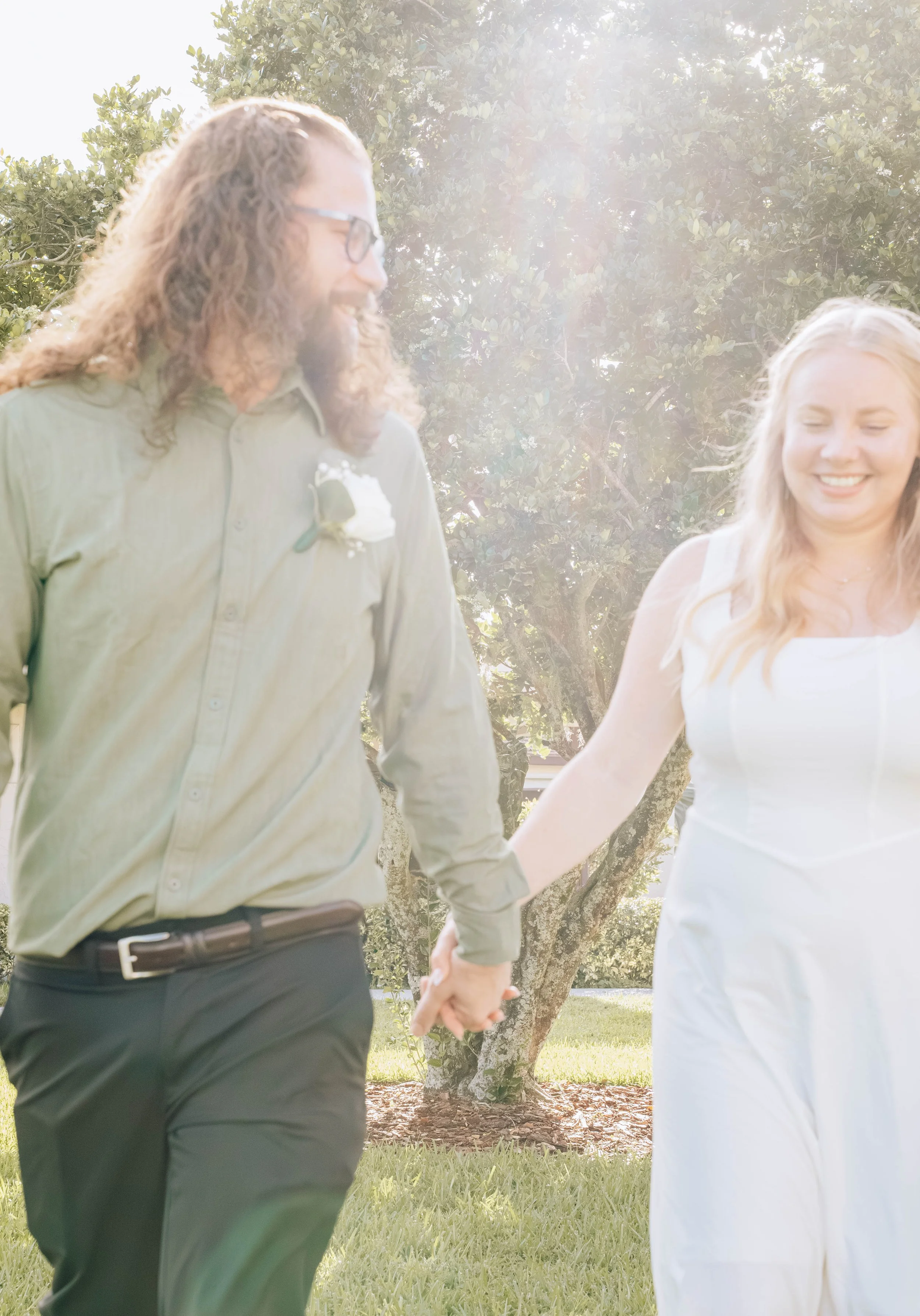 A couple holding hands at their wedding outdoors, with sunlight filtering through trees, both smiling and dressed in wedding attire.