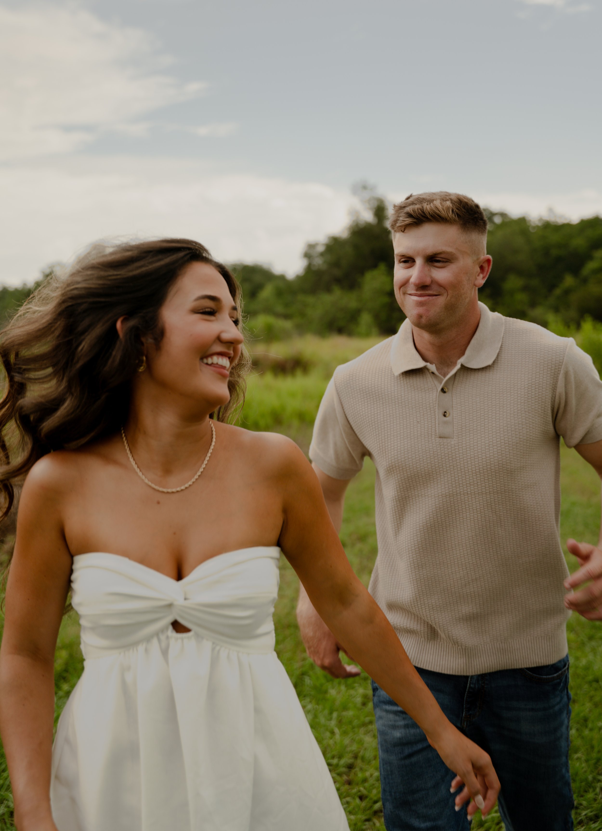 A woman in a white dress with a cutout is smiling and holding hands with a man outdoors in a grassy field, both appearing happy.