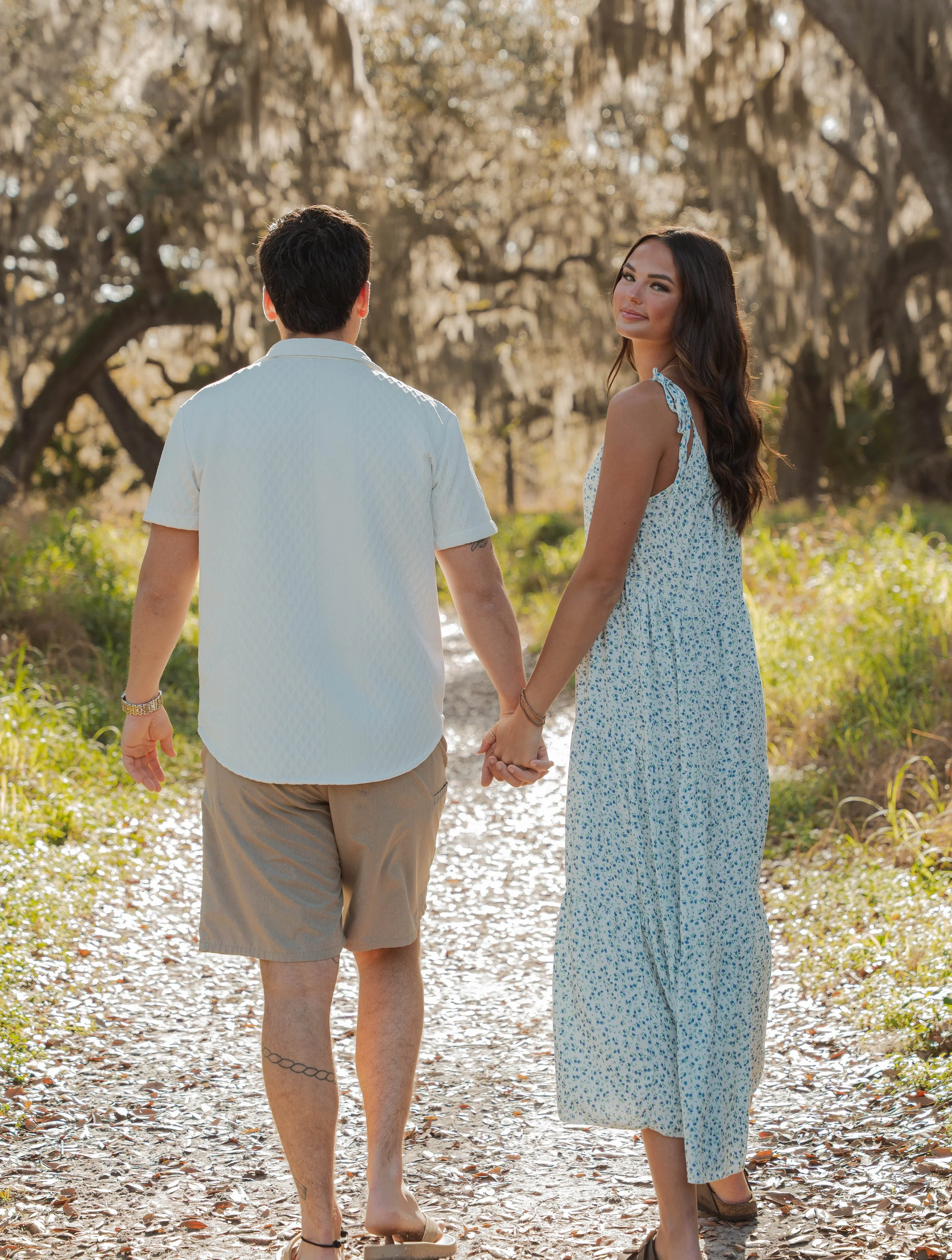 A couple holding hands walking on a forest path. The woman is smiling and looking back at the camera, while the man faces away. They are dressed in summer clothes, with the woman in a blue floral dress and the man in a white shirt and beige shorts.