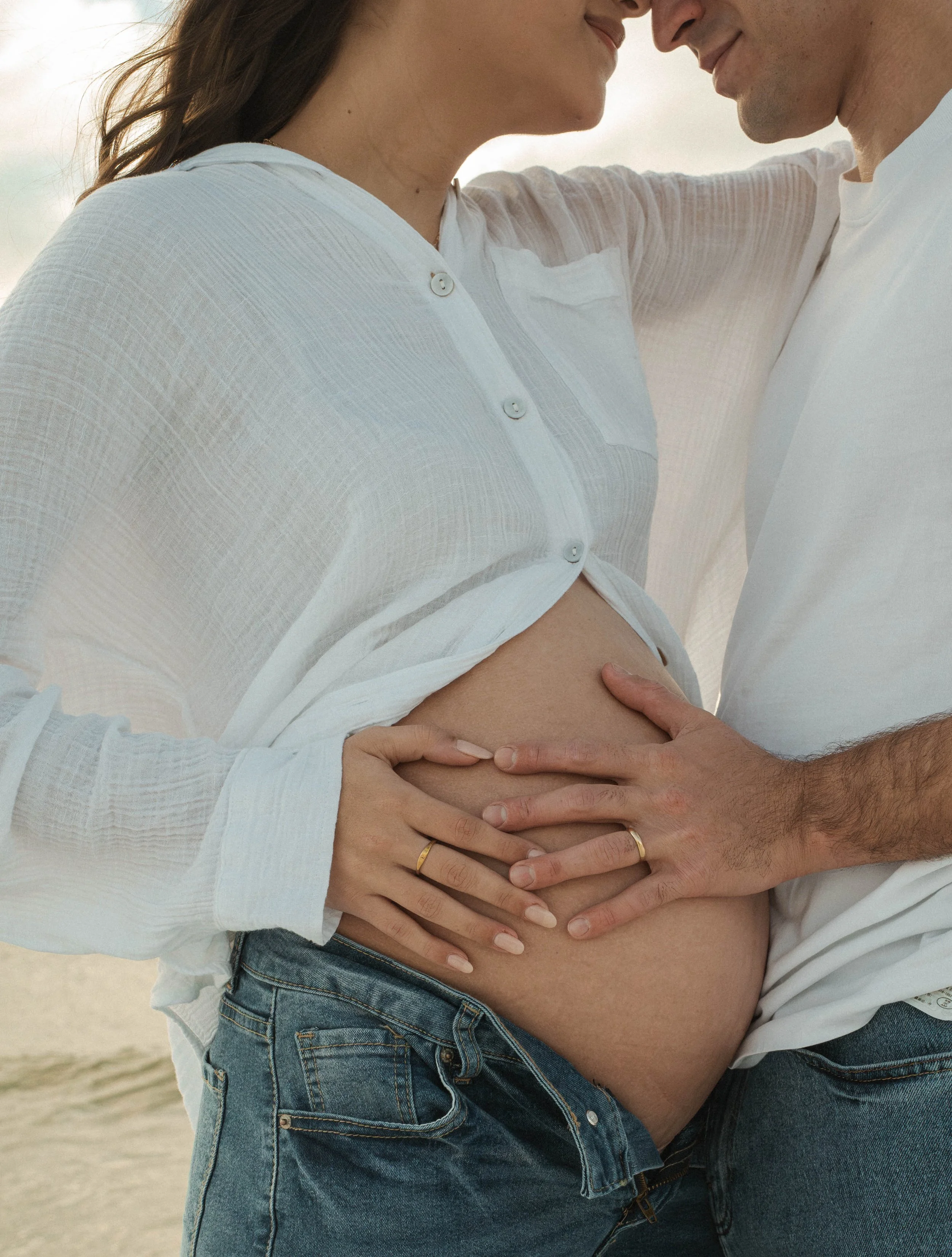 A pregnant woman and a man embrace, touching her belly, with their faces close together and smiling softly. The woman wears a white, semi-sheer shirt and jeans, while the man wears a white shirt.