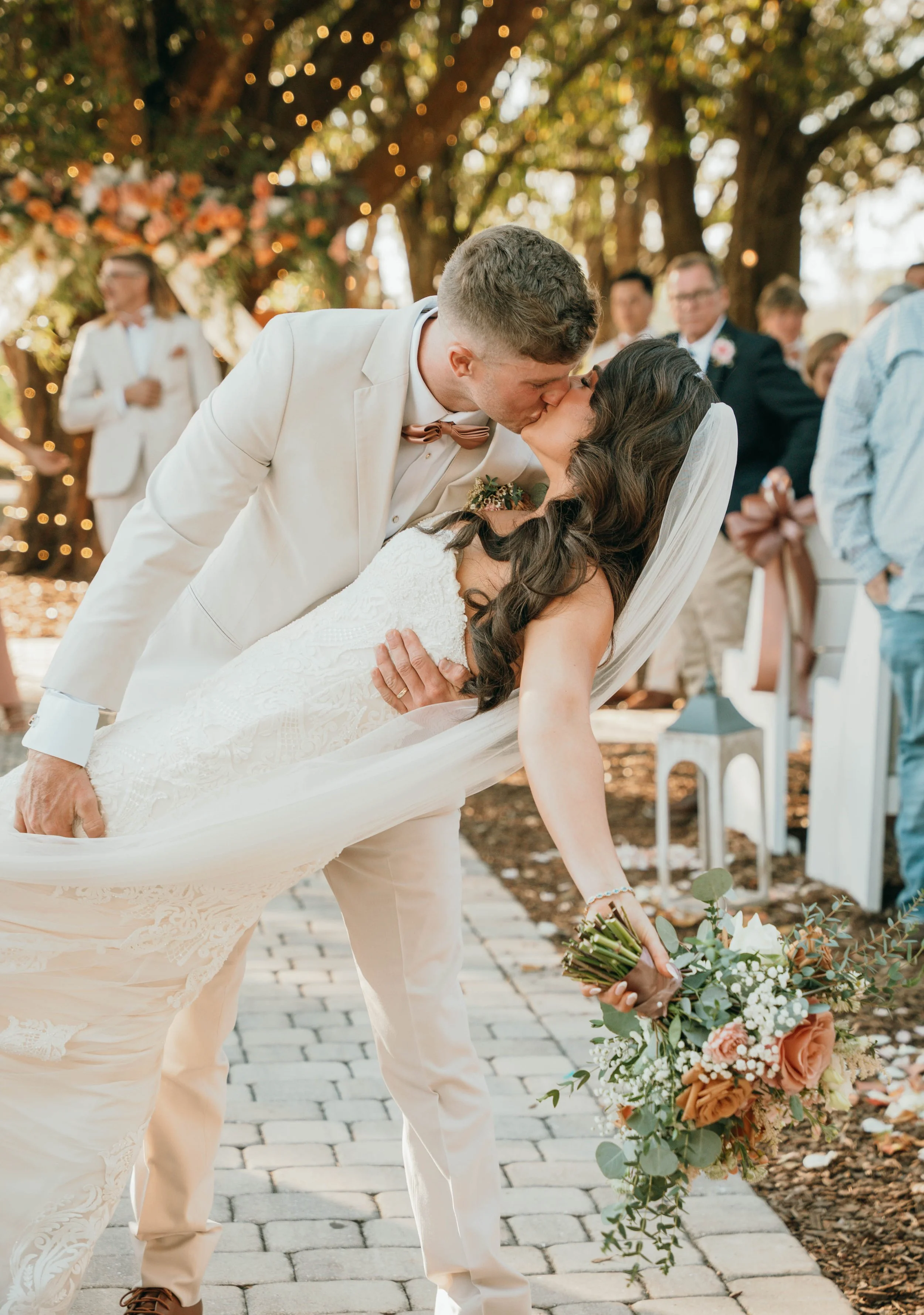 A newlywed couple sharing a kiss during their outdoor wedding ceremony in a garden decorated with string lights and flowers.