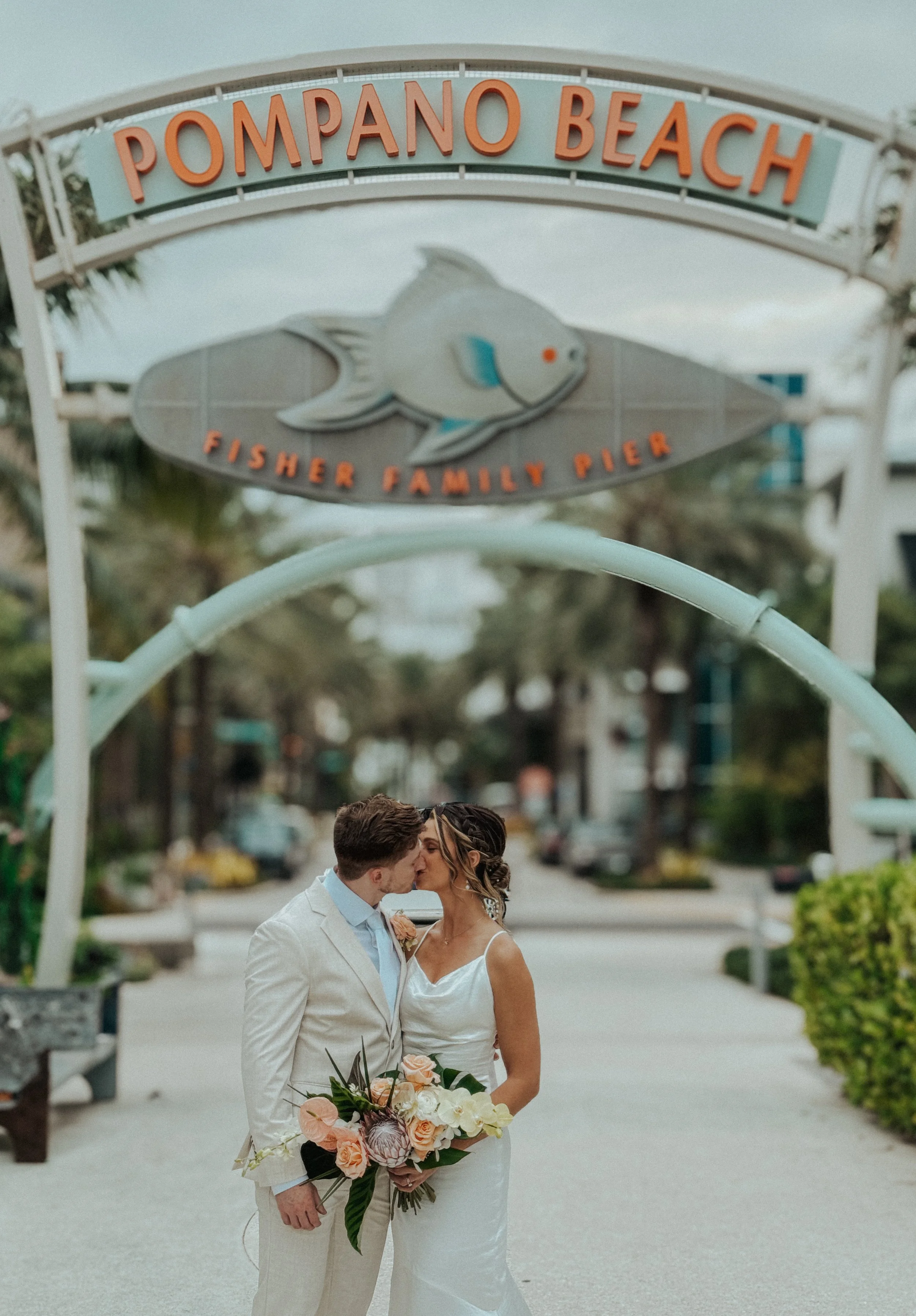 A newlywed couple is sharing a kiss under a sign that reads "Pompano Beach" and "Fisher Family Pier" while standing on a sidewalk with trees and buildings in the background.