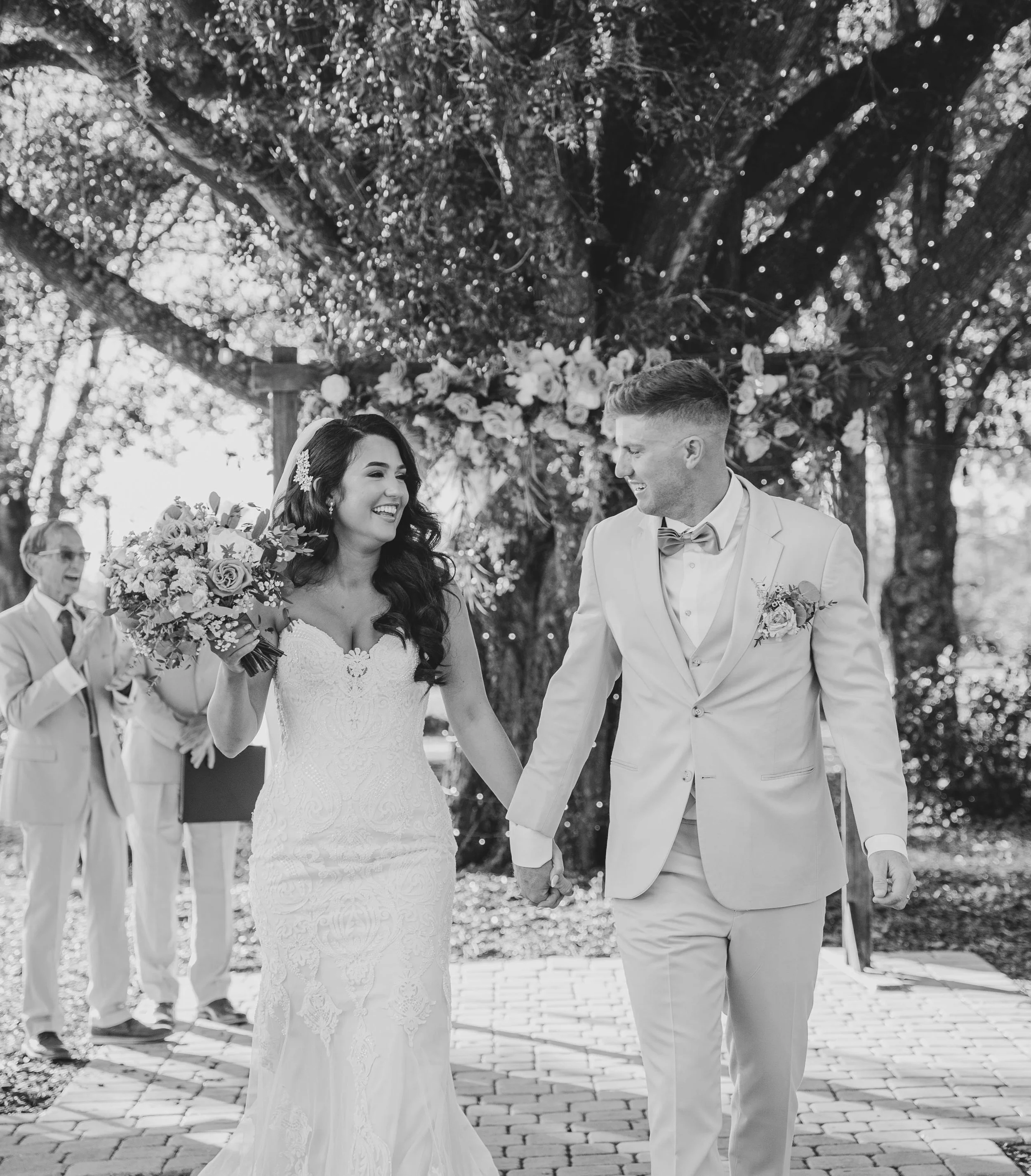 A black and white photo of a bride and groom holding hands and smiling at each other during their outdoor wedding ceremony. The bride is wearing a lace wedding dress and holding a bouquet, while the groom is dressed in a light-colored suit with a bow tie. There is floral decoration and a large tree in the background.