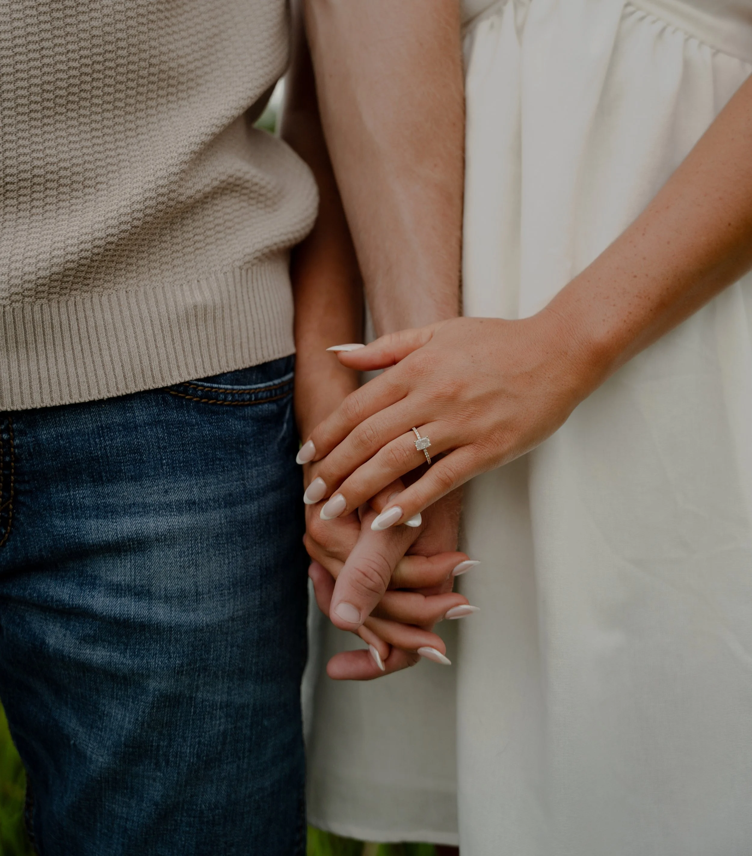 Close-up of a couple holding hands, with a focus on an engagement ring on the woman's finger, showing the woman in a white dress and the man in a beige sweater and blue jeans.