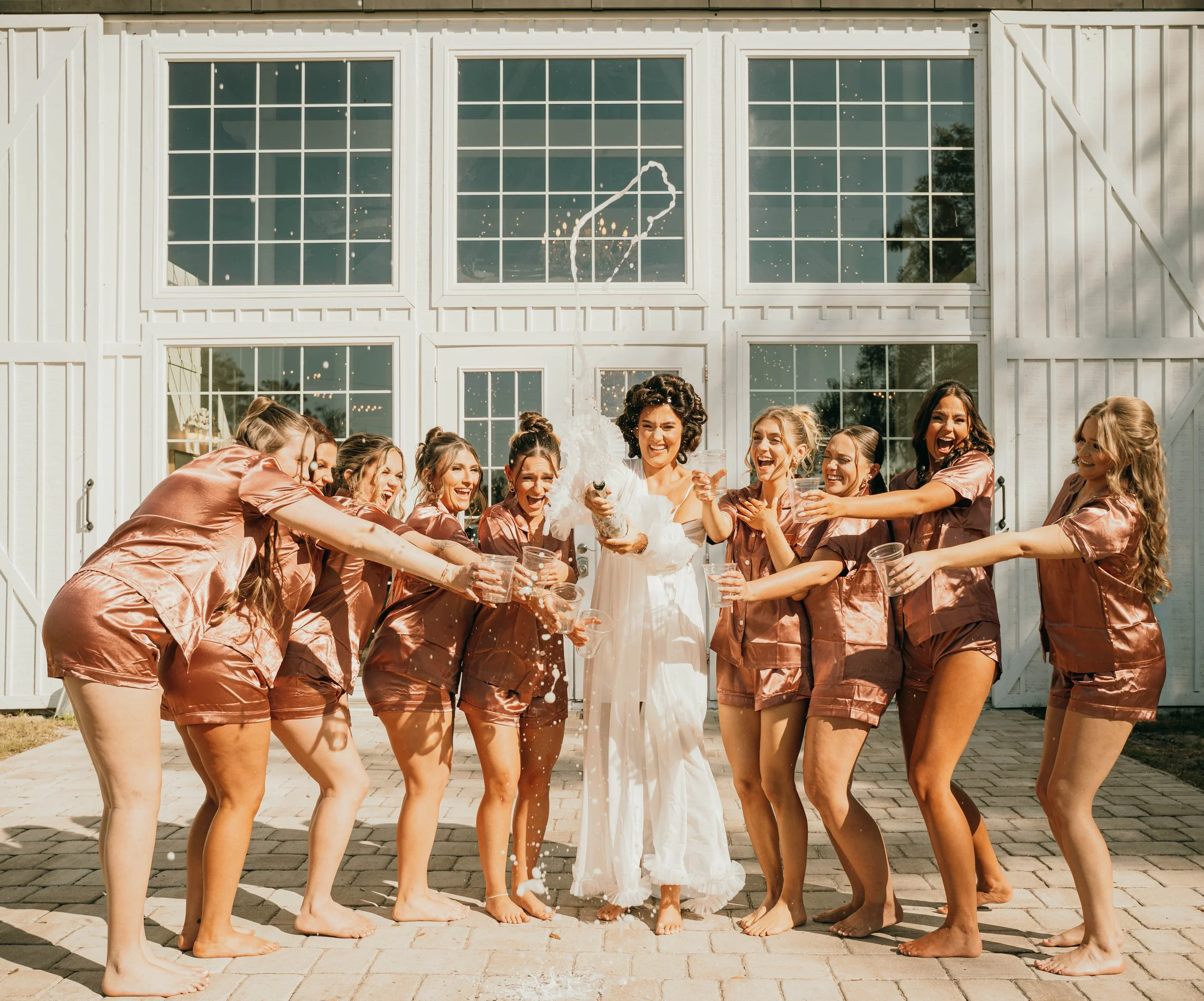 Bride in white dress celebrating with bridesmaids in matching satin pajamas outside a white barn, having a cake smash with drinks.