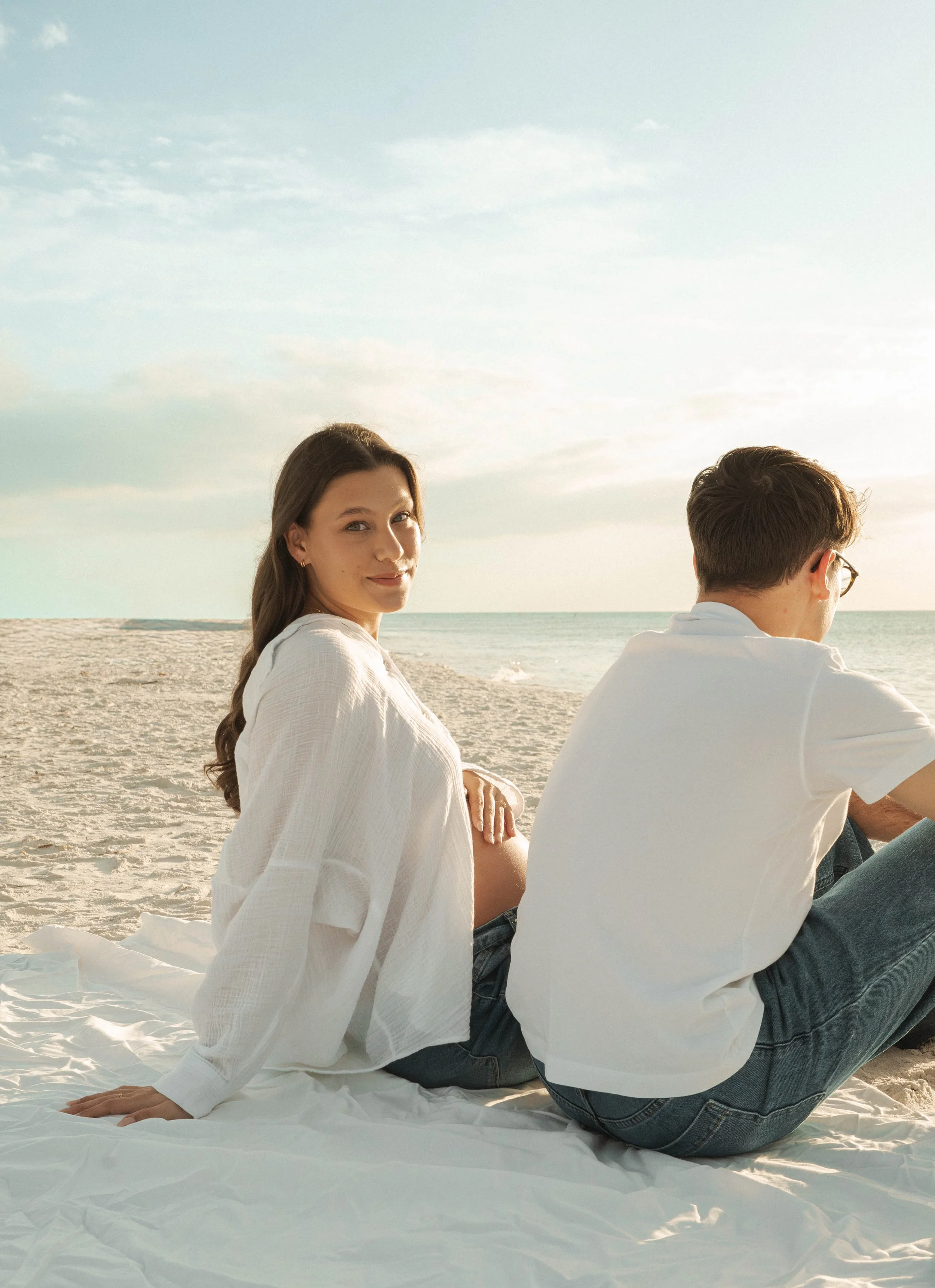 A beautiful pregnant woman and her husband sitting on a white blanket on a sandy beach during sunset, with the woman facing the camera and the man facing away, near the ocean.