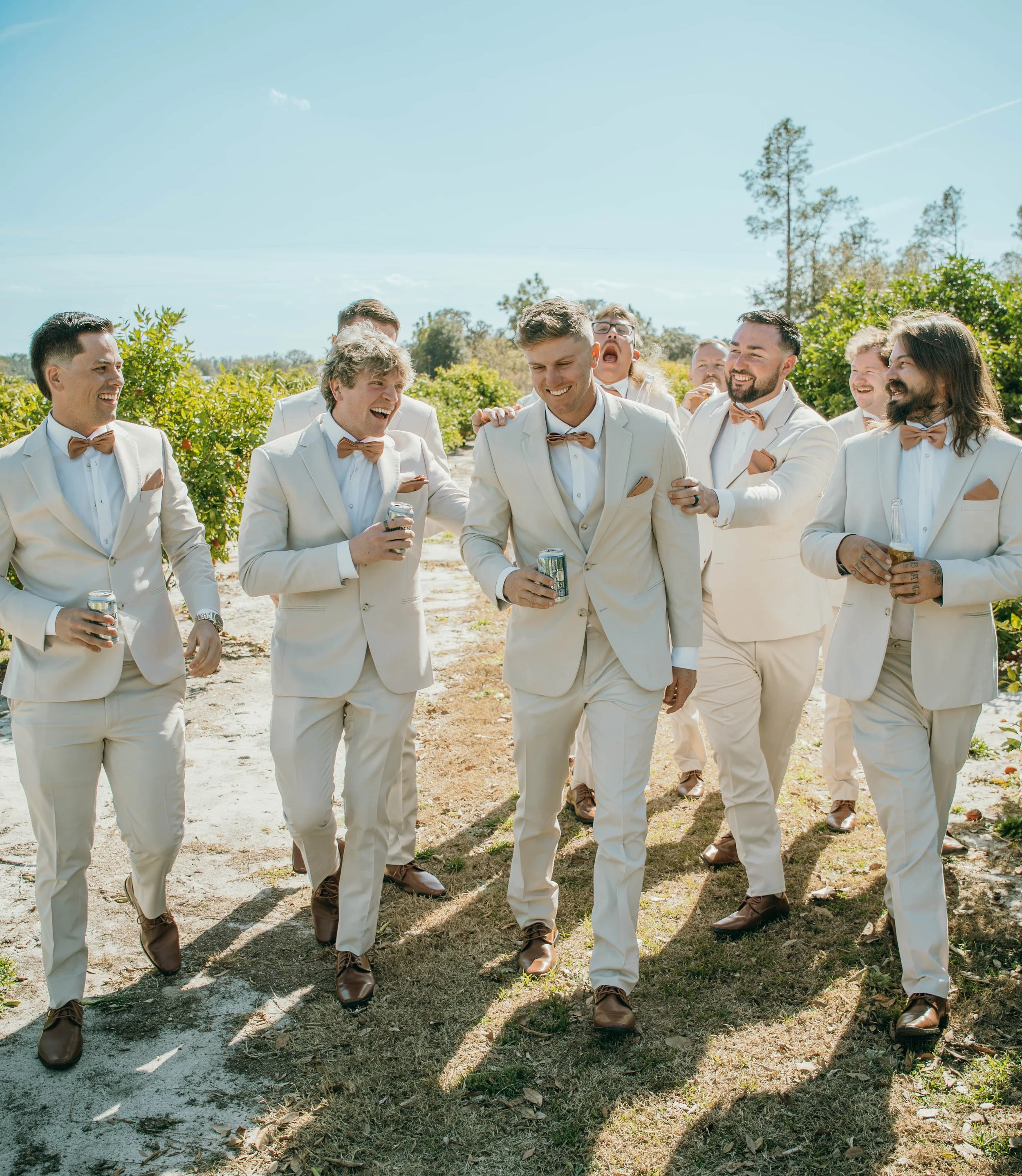 Groomsmen in cream tuxedos with brown shoes and bow ties walking outdoors, holding drinks, smiling and chatting on a sunny day.