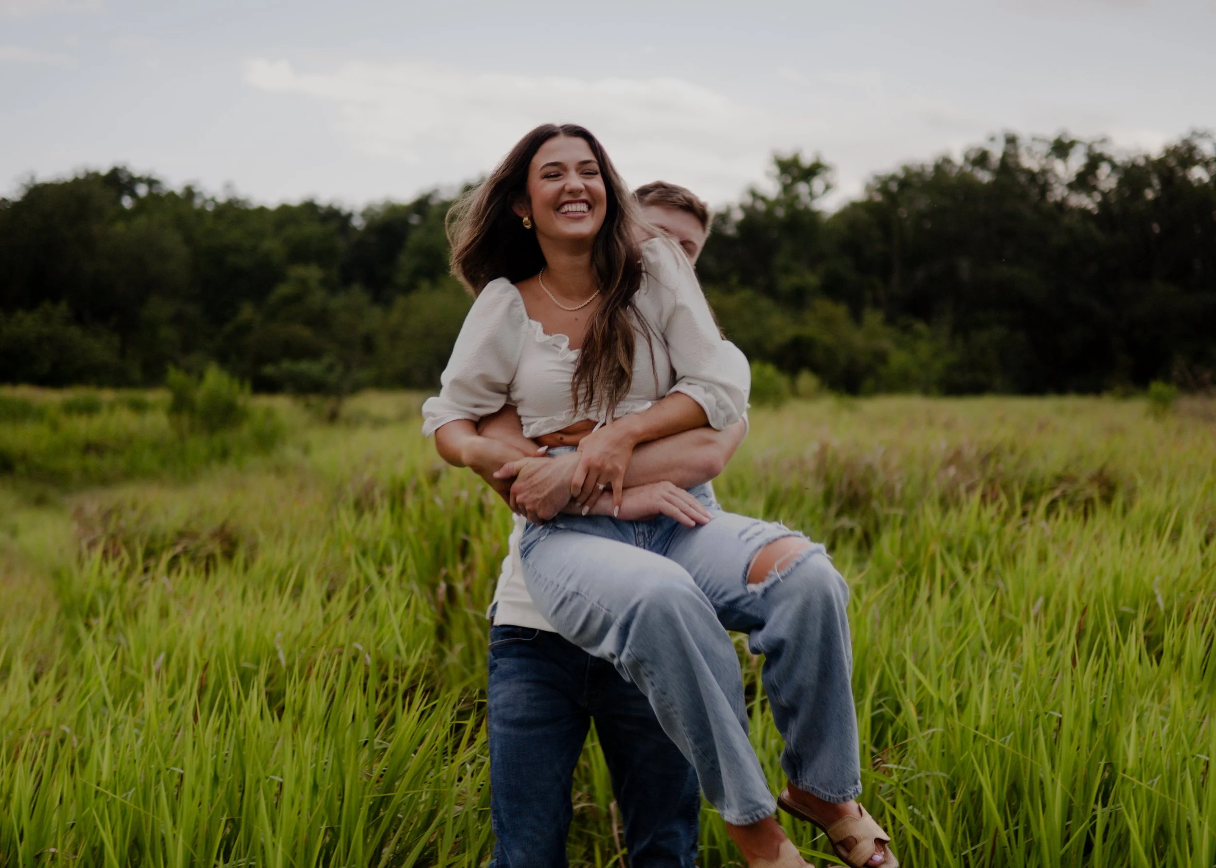 A woman in a white blouse and ripped jeans sitting on a man in a blue shirt and jeans, in a grassy field with trees in the background, during daytime.