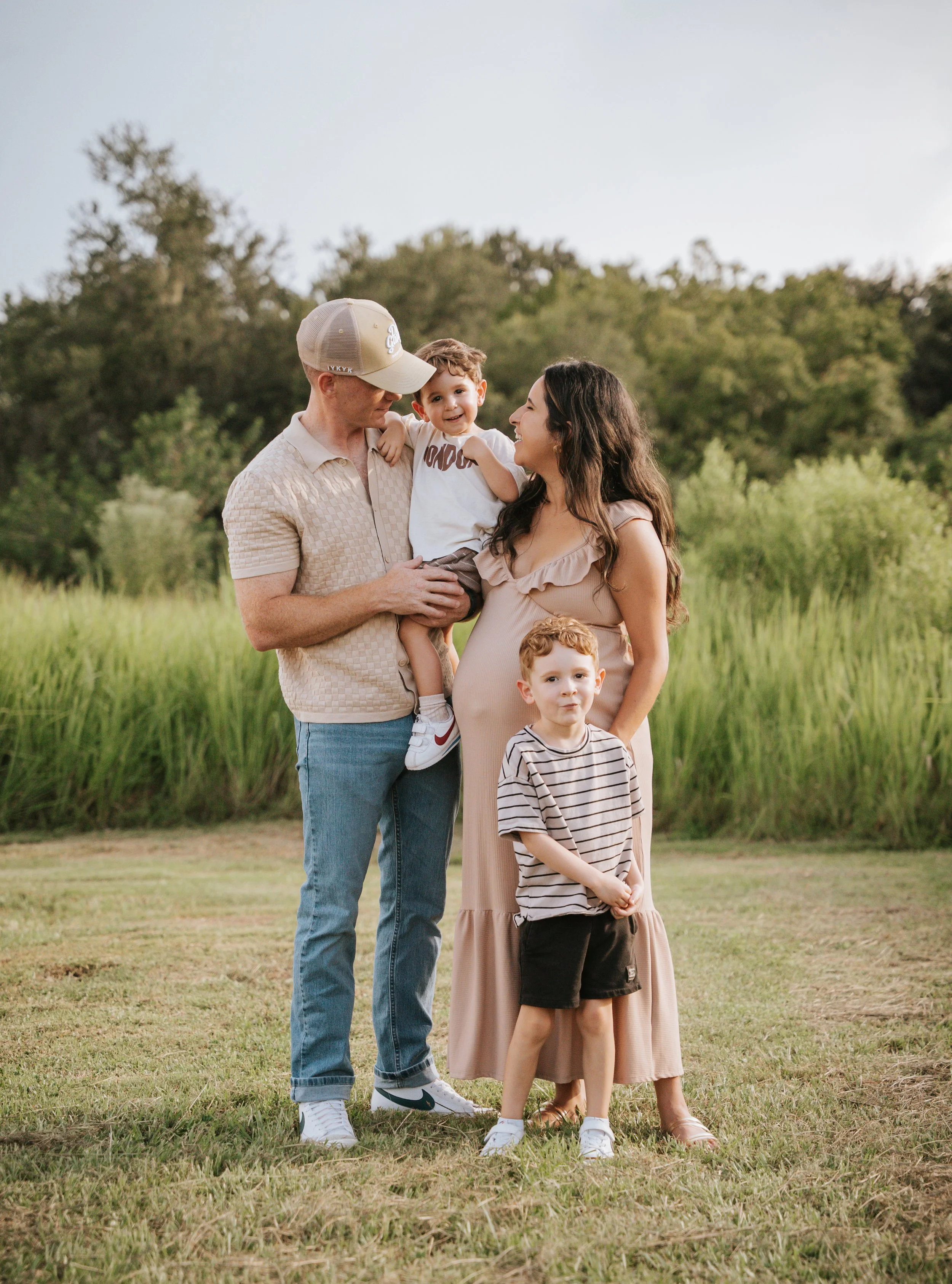 A family of four enjoying a moment outdoors in a grassy field with trees in the background. The father is holding a young boy, and the mother, who is pregnant, stands next to a boy in a striped shirt. They are all looking at each other and smiling.