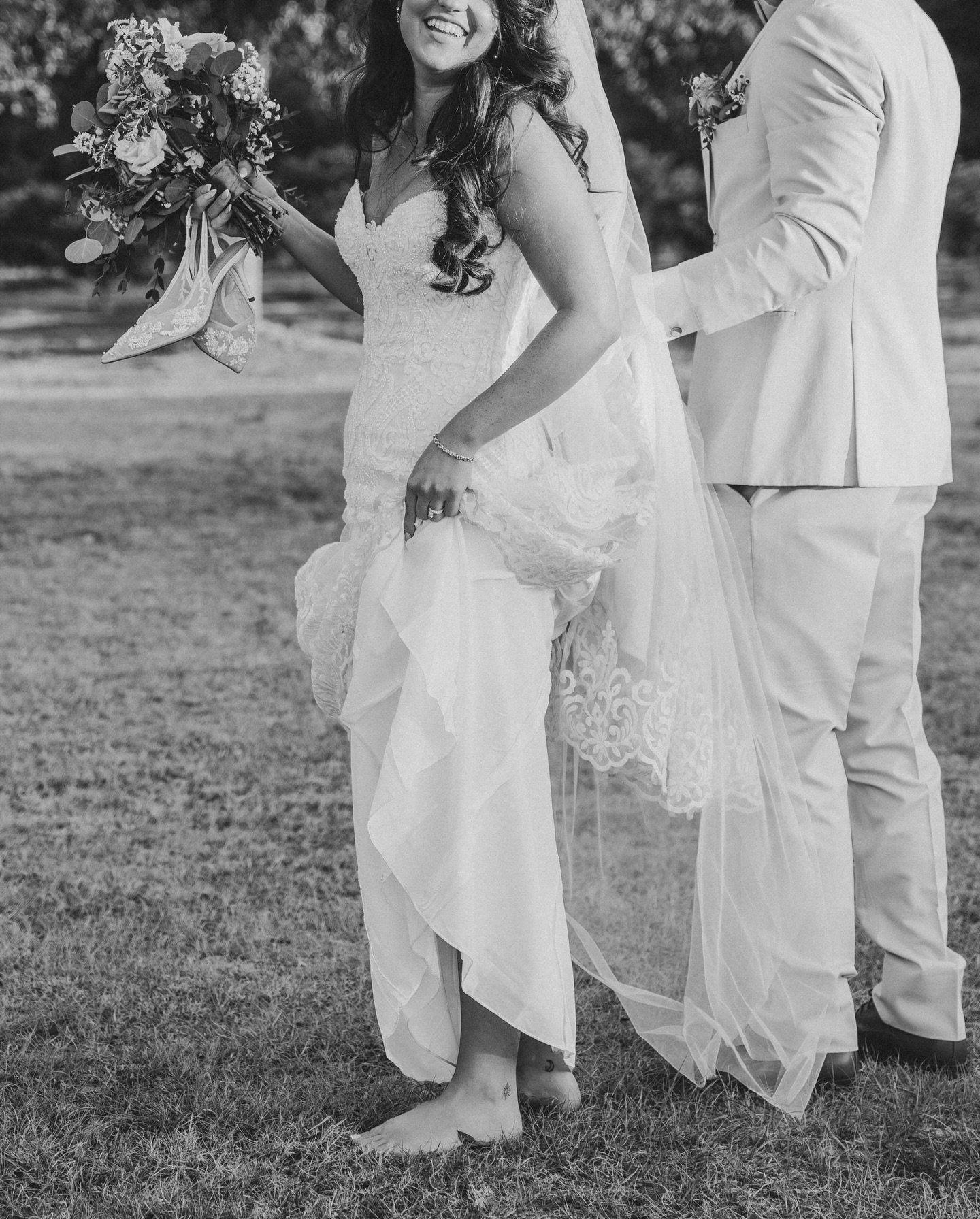 Black and white photo of a bride in a wedding dress holding a bouquet, lifting her dress with bare feet, and smiling. A groom in a light suit stands beside her, holding her waist.