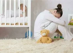 A sad woman sitting on the floor beside a crib with a baby inside, surrounded by toys including a teddy bear.