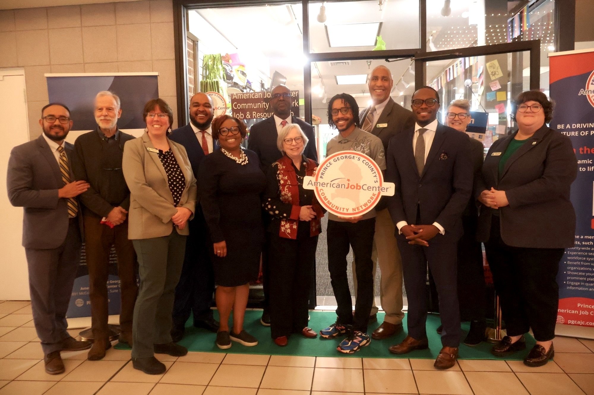 Group of diverse people standing together inside a building, holding a sign for Prince George's County American Job Center Community Network, smiling for a photo.