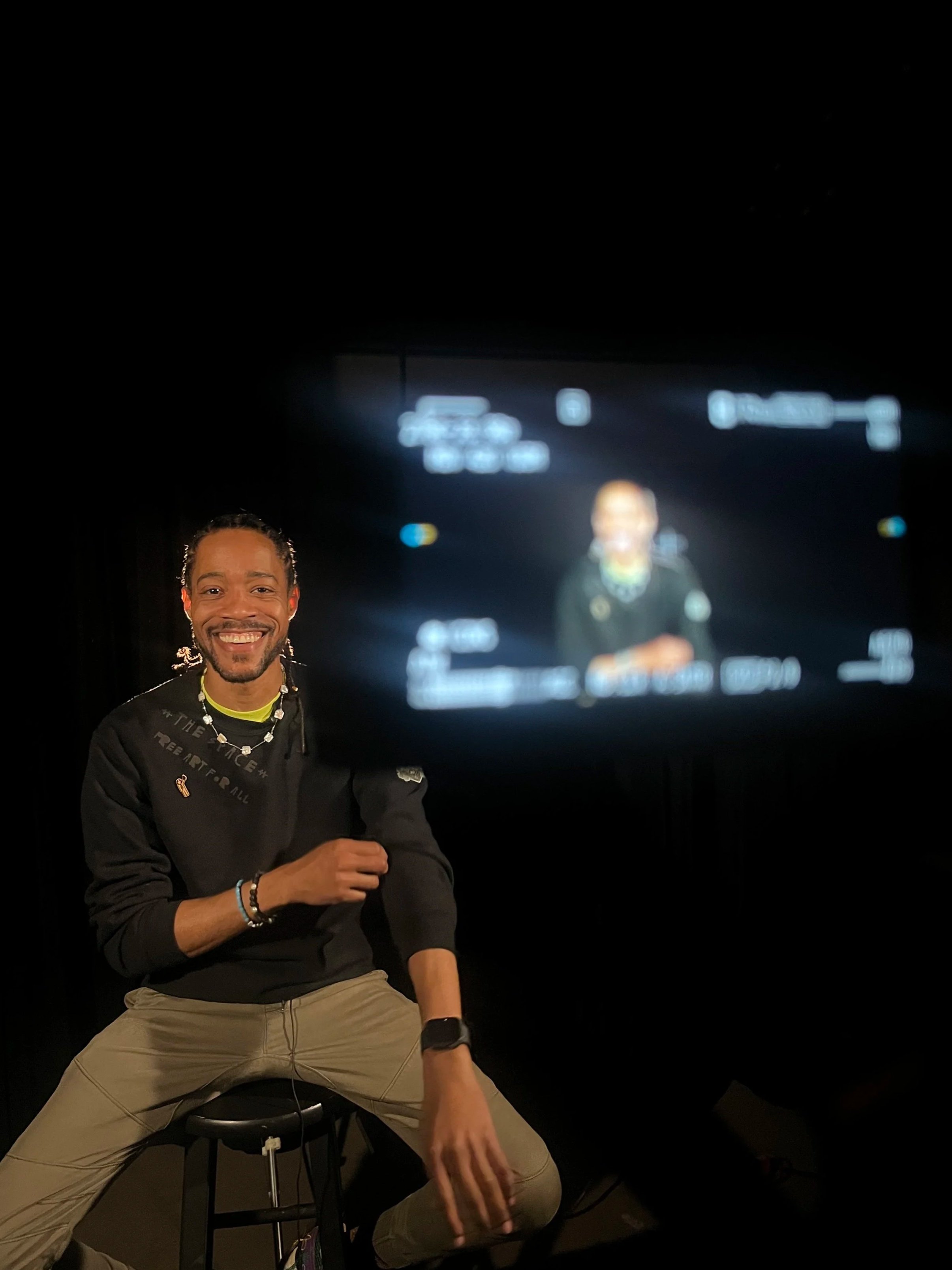 Man smiling and sitting on a stool in front of a camera monitor with a blurred image of himself in the background.