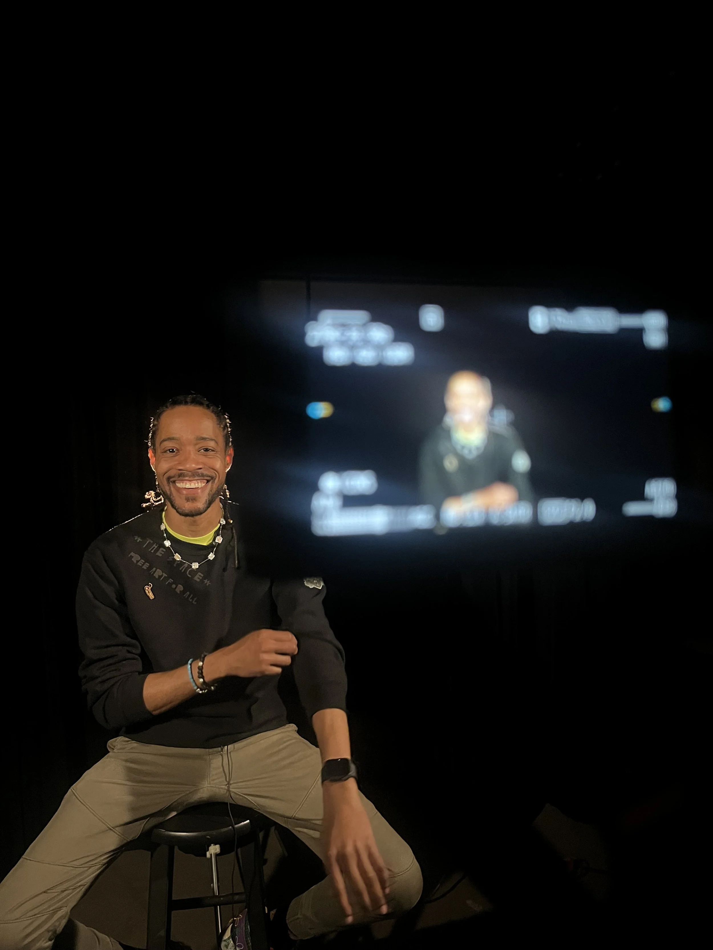 Smiling man sitting on a stool in a dark room with a large camera screen behind him, capturing his photo.