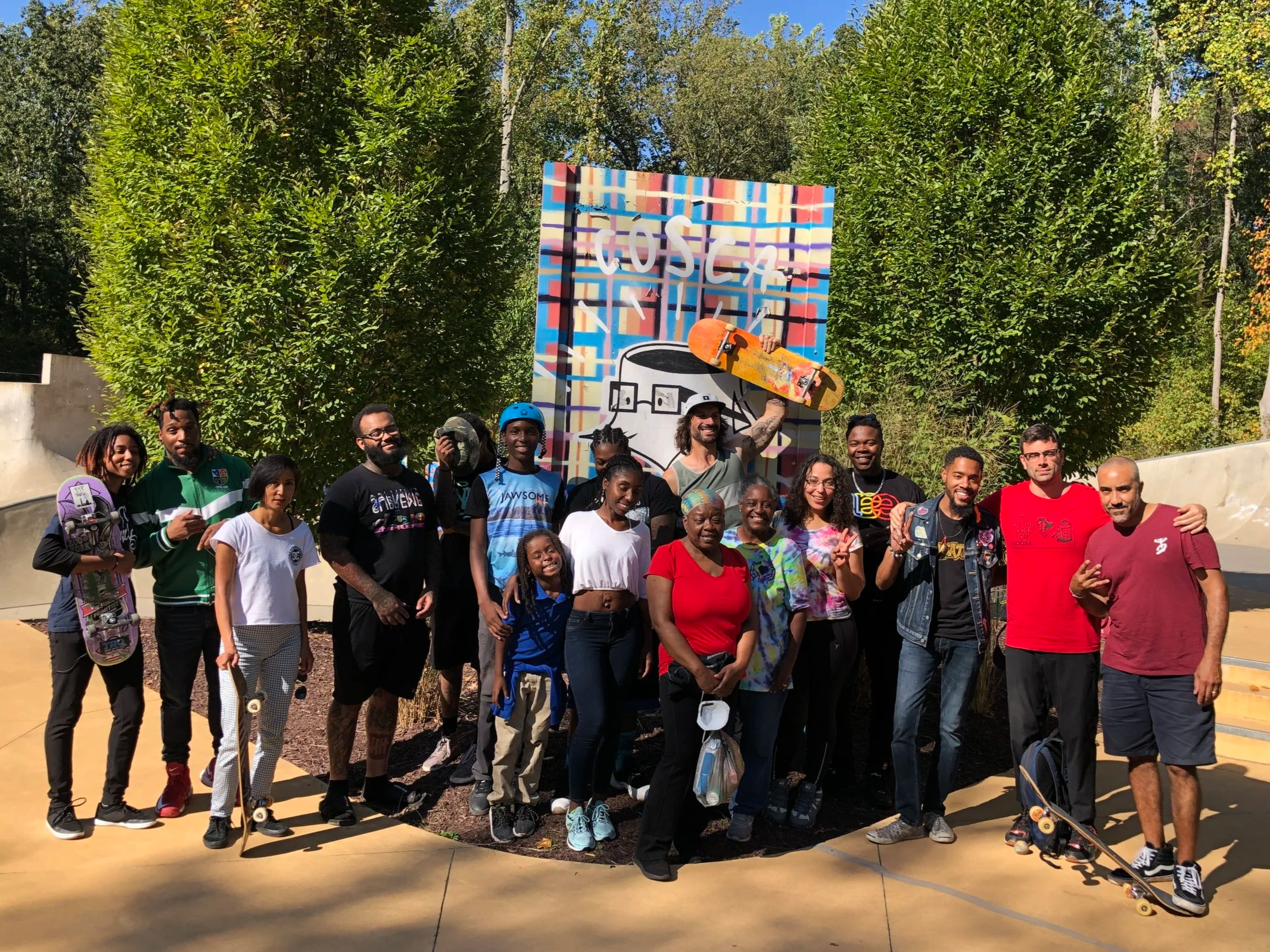 Group of diverse people at a skate park, smiling and holding skateboards, with a colorful graffiti wall and large green trees in the background.