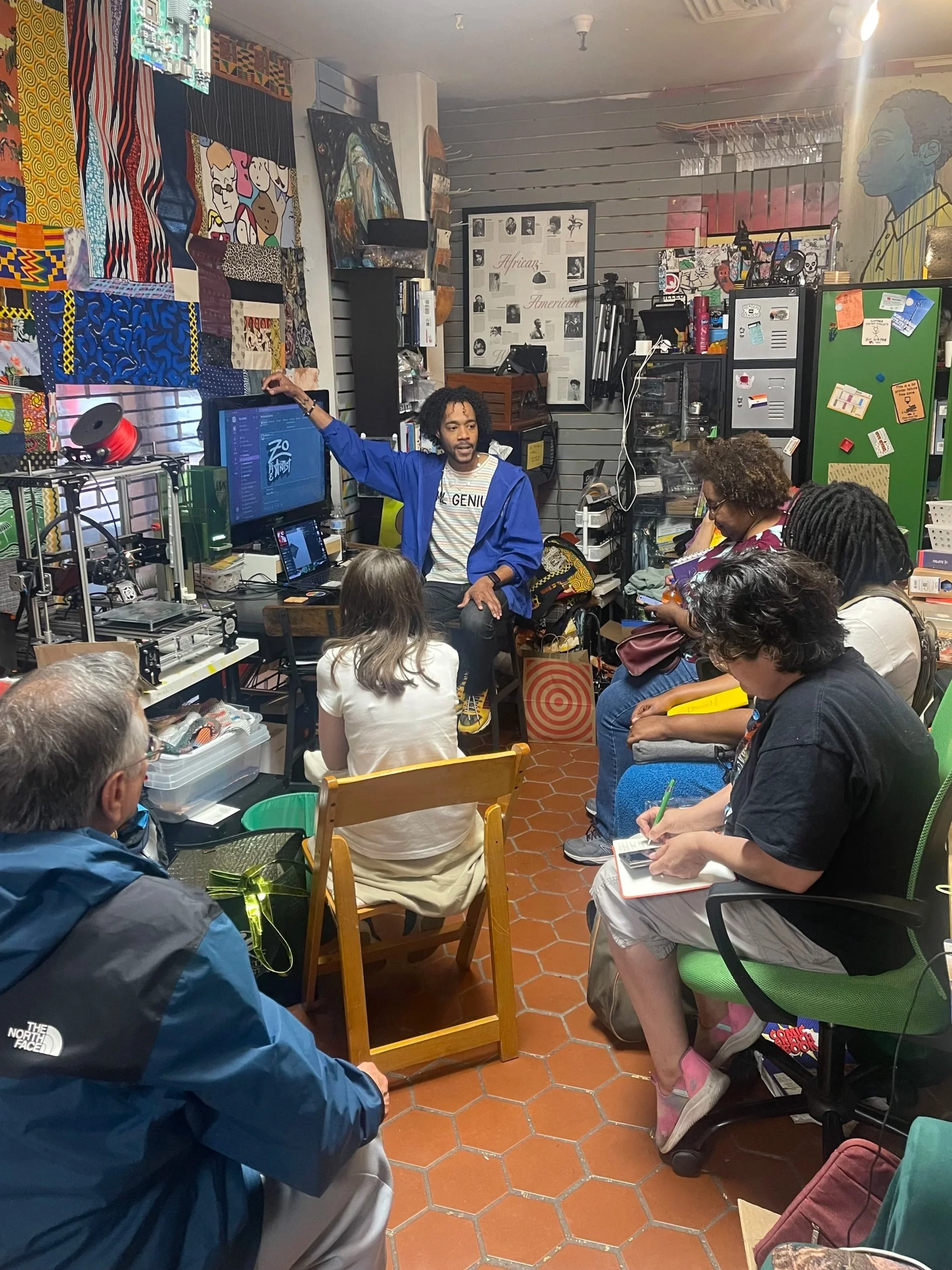 A man in a blue jacket is giving a presentation to a small audience seated in a cluttered room decorated with artwork, fabric, and posters.