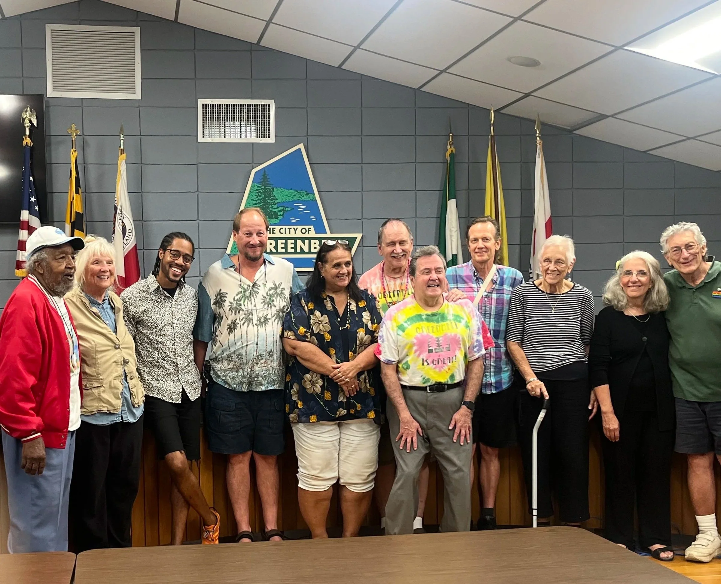 Group of diverse people standing together at a city hall, smiling, with flags and a city emblem in the background, celebrating an event.