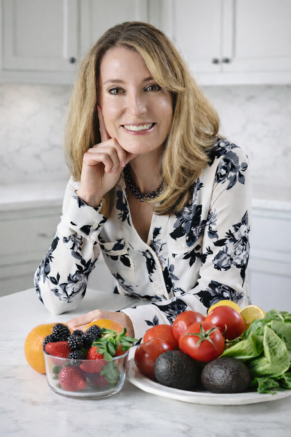 A woman smiling at the camera with a hand resting on her chin in a kitchen. In front of her on the table are fresh vegetables and fruits including tomatoes, avocados, lettuce, strawberries, blackberries, and oranges.