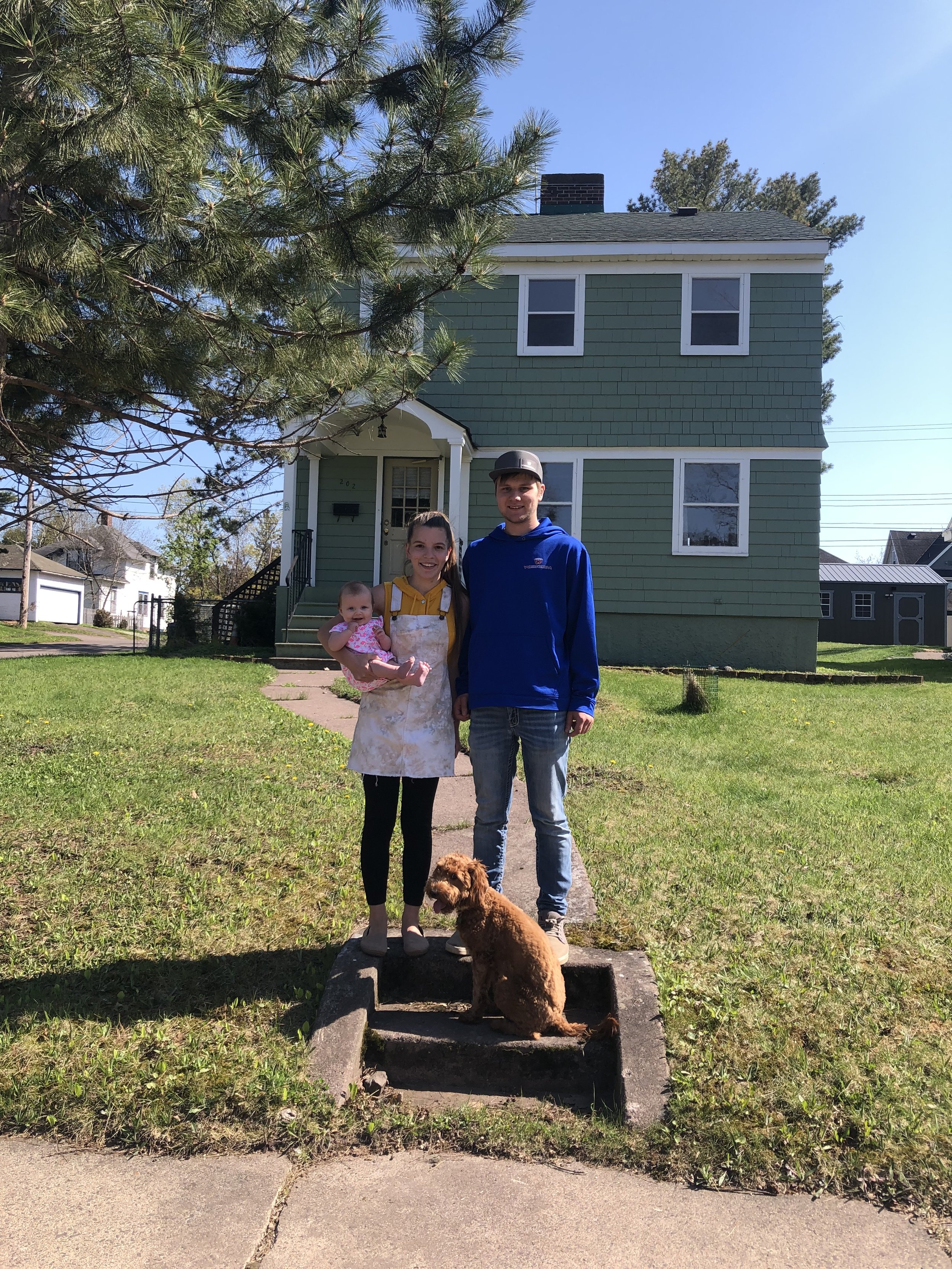 A family of three with a dog standing on front steps of a house. The woman is holding a baby, all are smiling. The house is green with white trim, and there is a large tree on the left side of the image. Clear blue sky in the background.