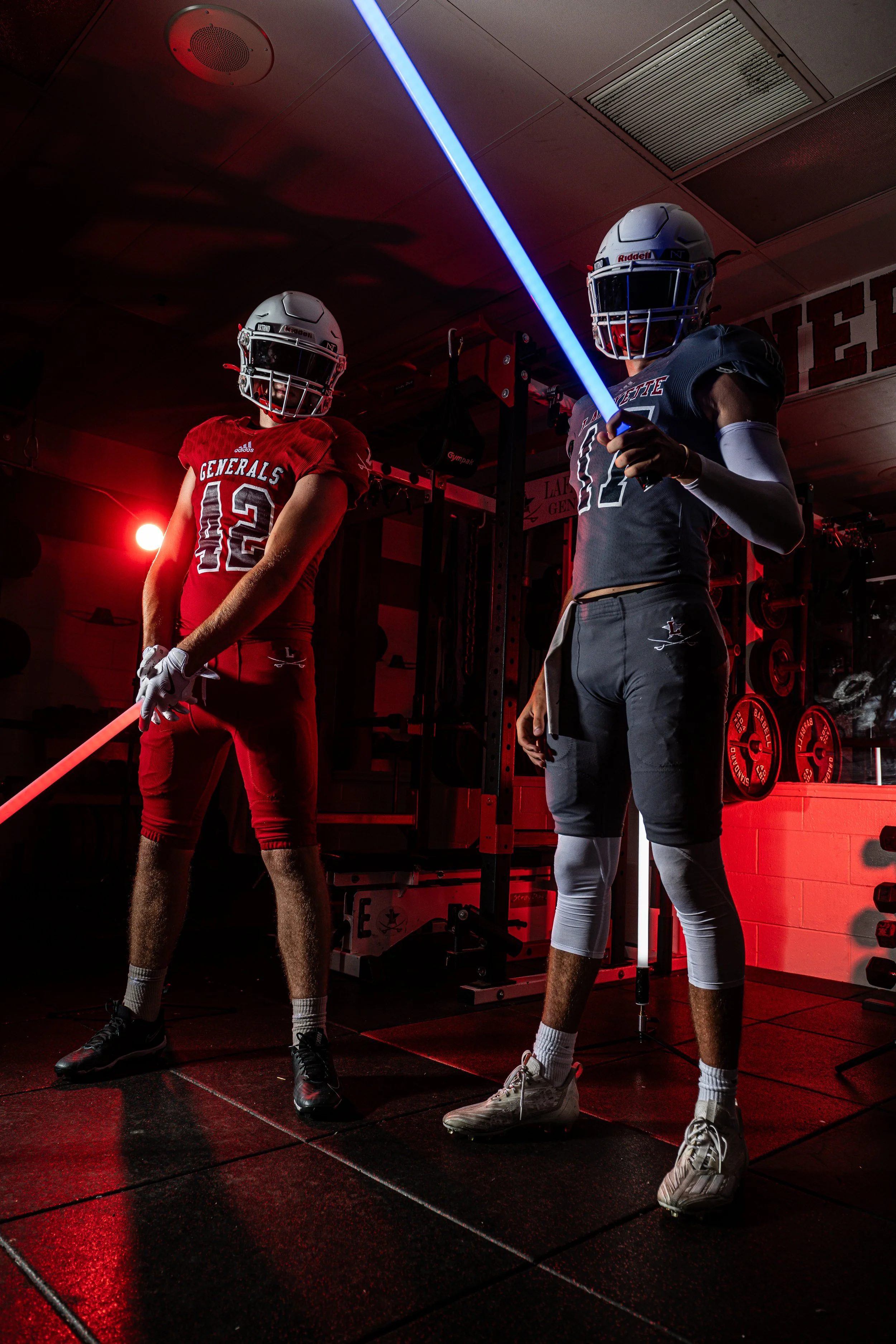Two football players in helmets and uniforms stand in a gym, with one holding a blue LED light saber. The gym is dimly lit with red and blue lighting.