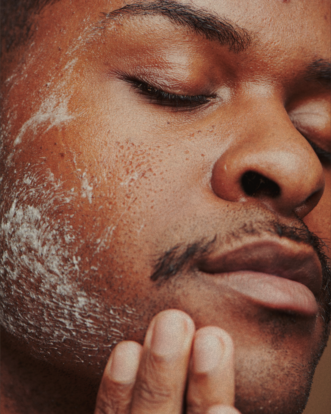 Close-up of a man's face with his eyes closed, applying a white facial scrub or mask to his cheek with his fingers.