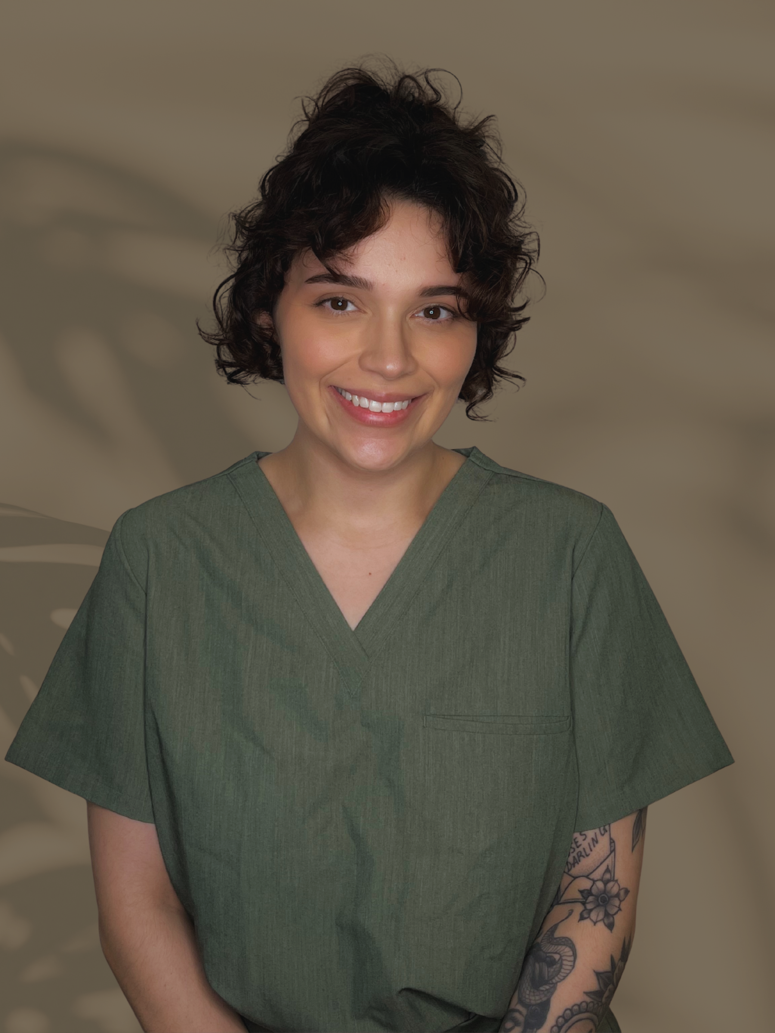 A young woman with short, curly brown hair and light skin, smiling, wearing green medical scrubs, with tattoos on her left arm, standing in front of a blurred neutral-toned background.