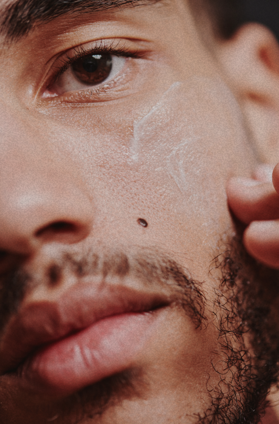 Close-up of a man's face with a skincare product on his cheek, showing his eye, nose, lips, facial hair, and a mole.
