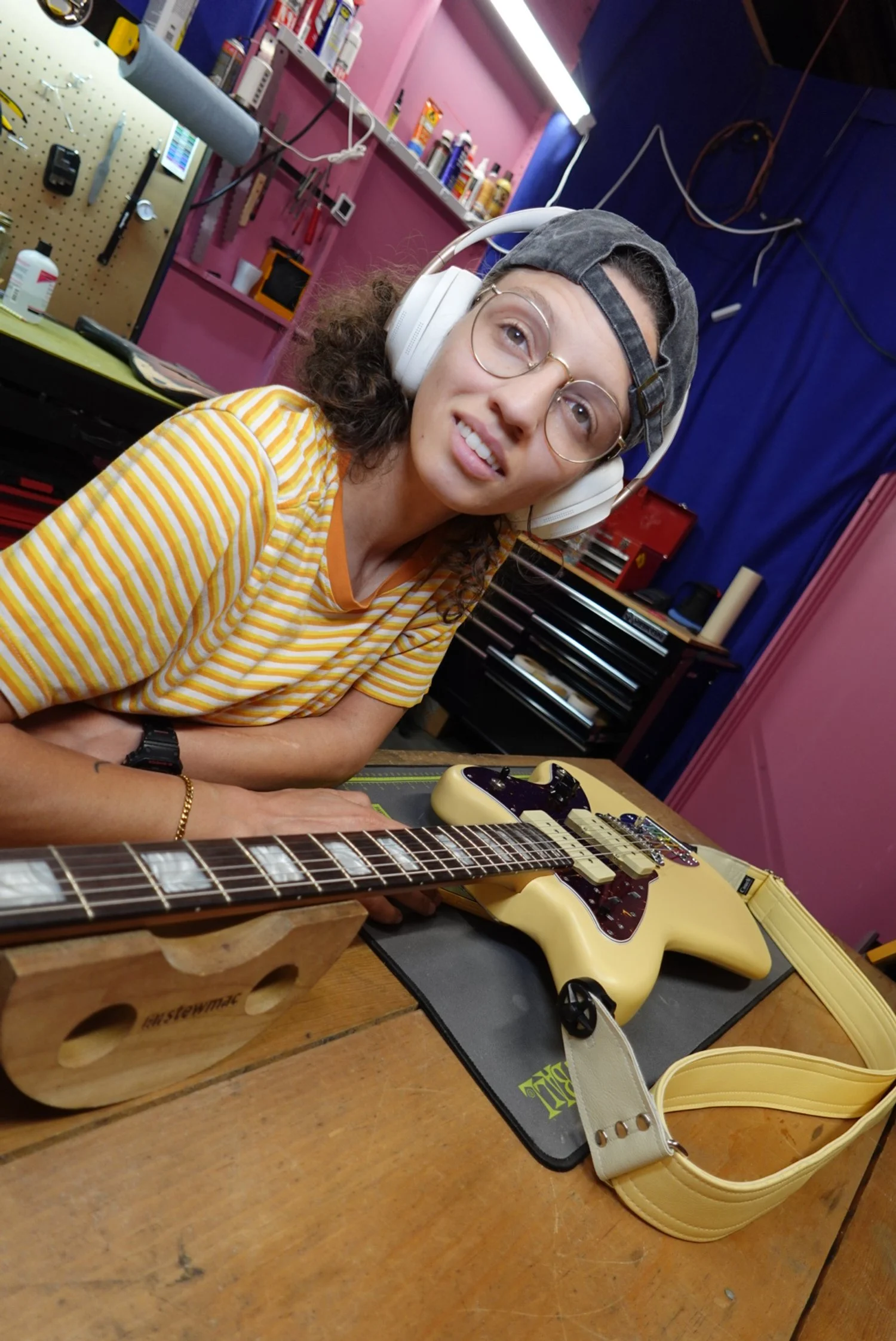 A person with curly hair, glasses, and a backwards cap wears headphones and a yellow striped shirt, sitting at a workshop table with a yellow electric guitar in front of them.