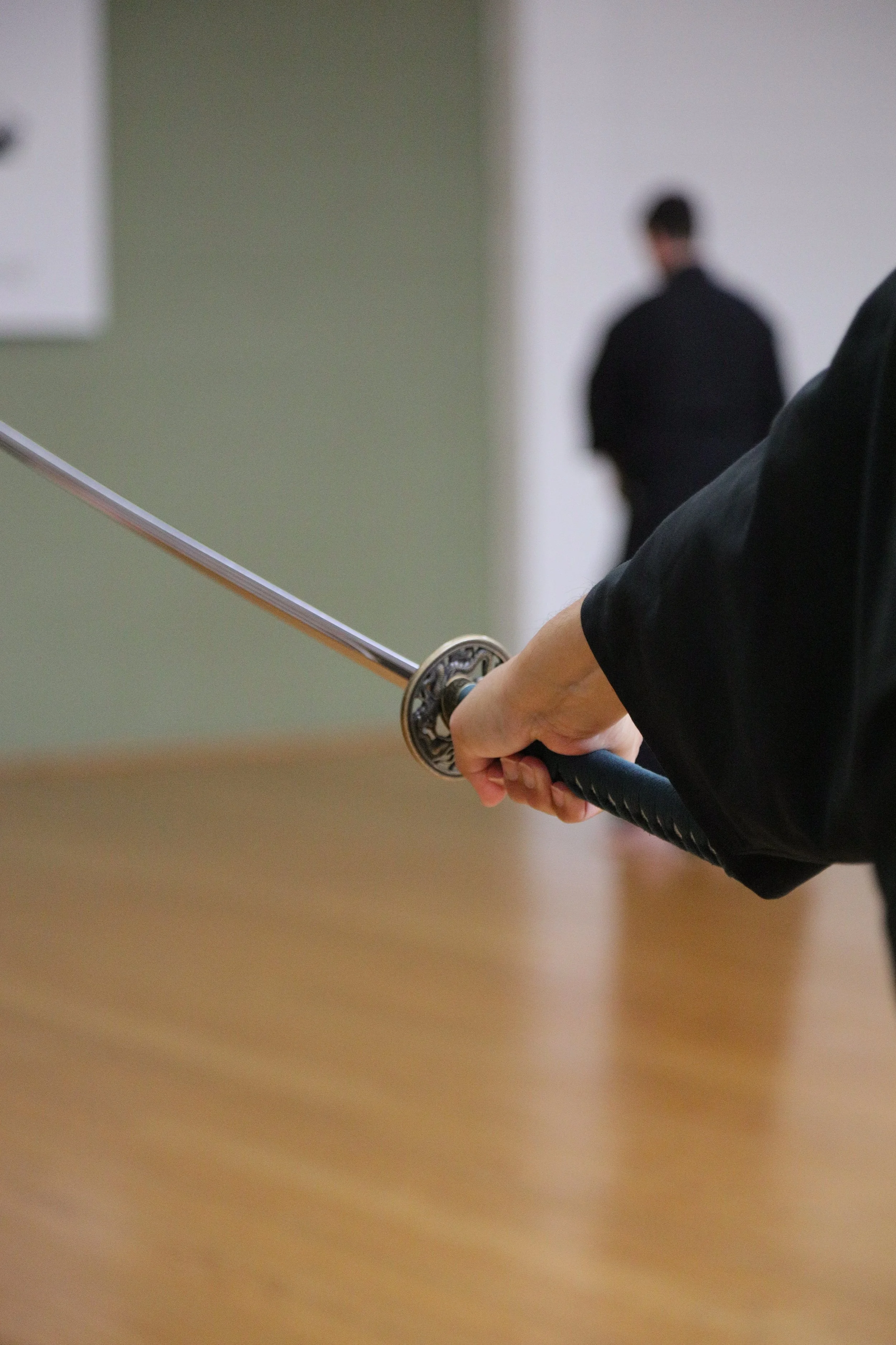 Close-up of a person's hand holding a katana sword, with the blade extended. In the background, a person dressed in black stands in a modern indoor space, slightly out of focus.